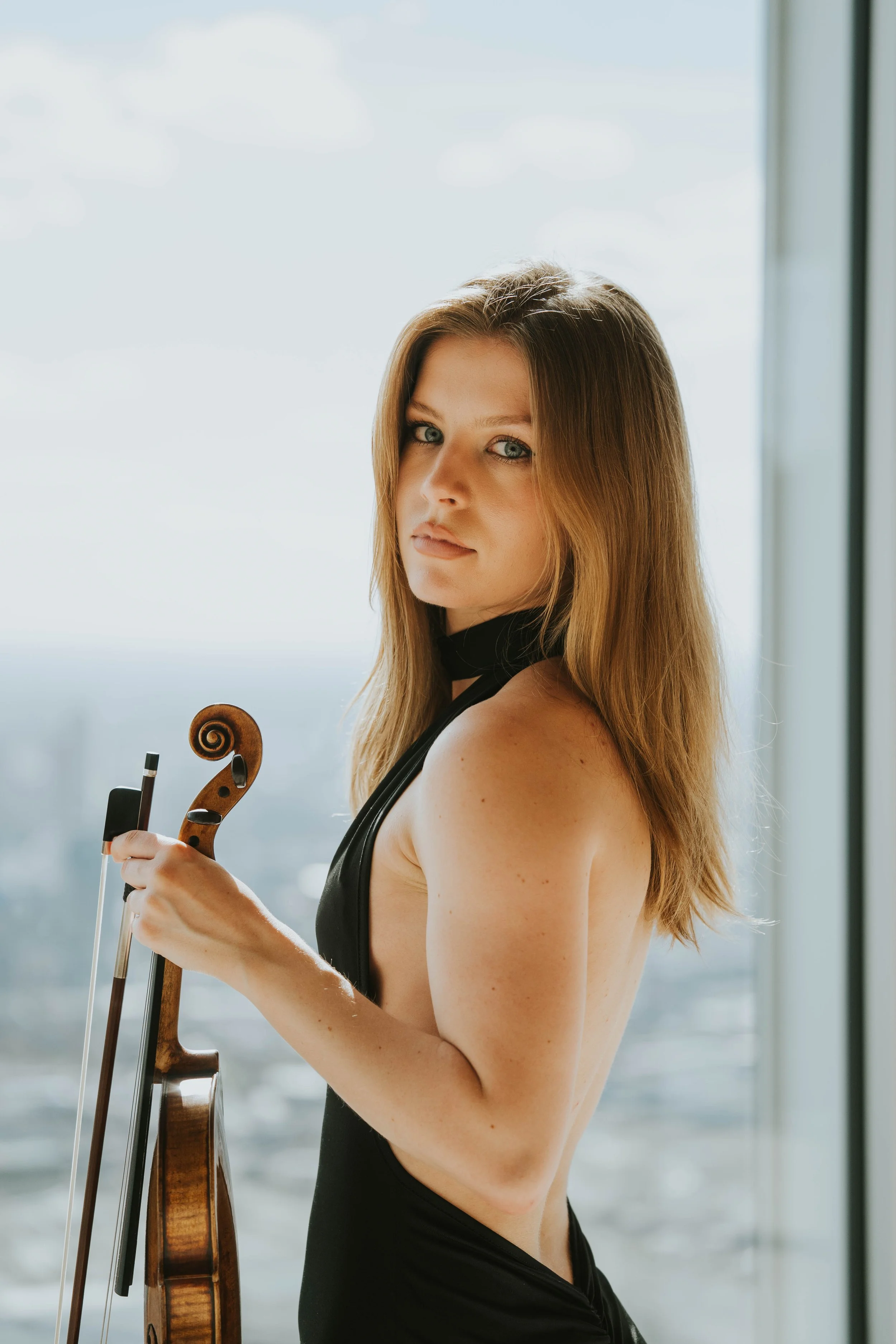A woman with long, light brown hair and blue eyes is holding a violin and bow, standing near a large window with a city view in the background.