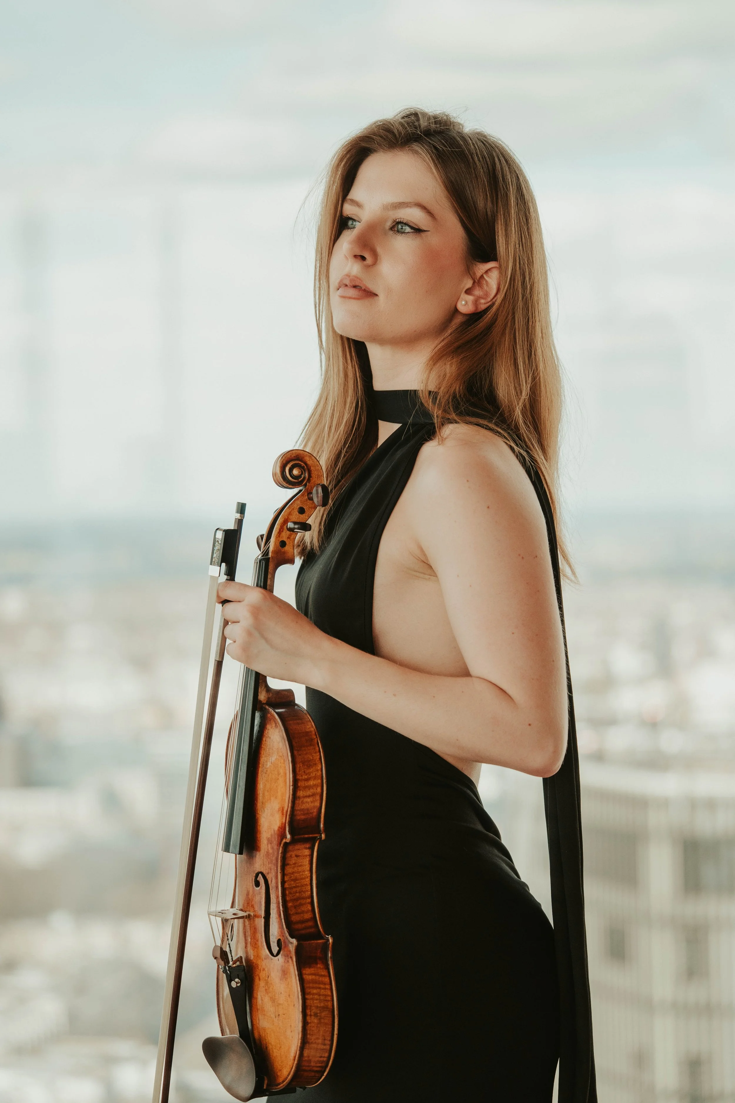 A woman with long brown hair and blue eyes wearing a black dress, holding a violin and bow, standing in front of a large window with a bright cityscape in the background.