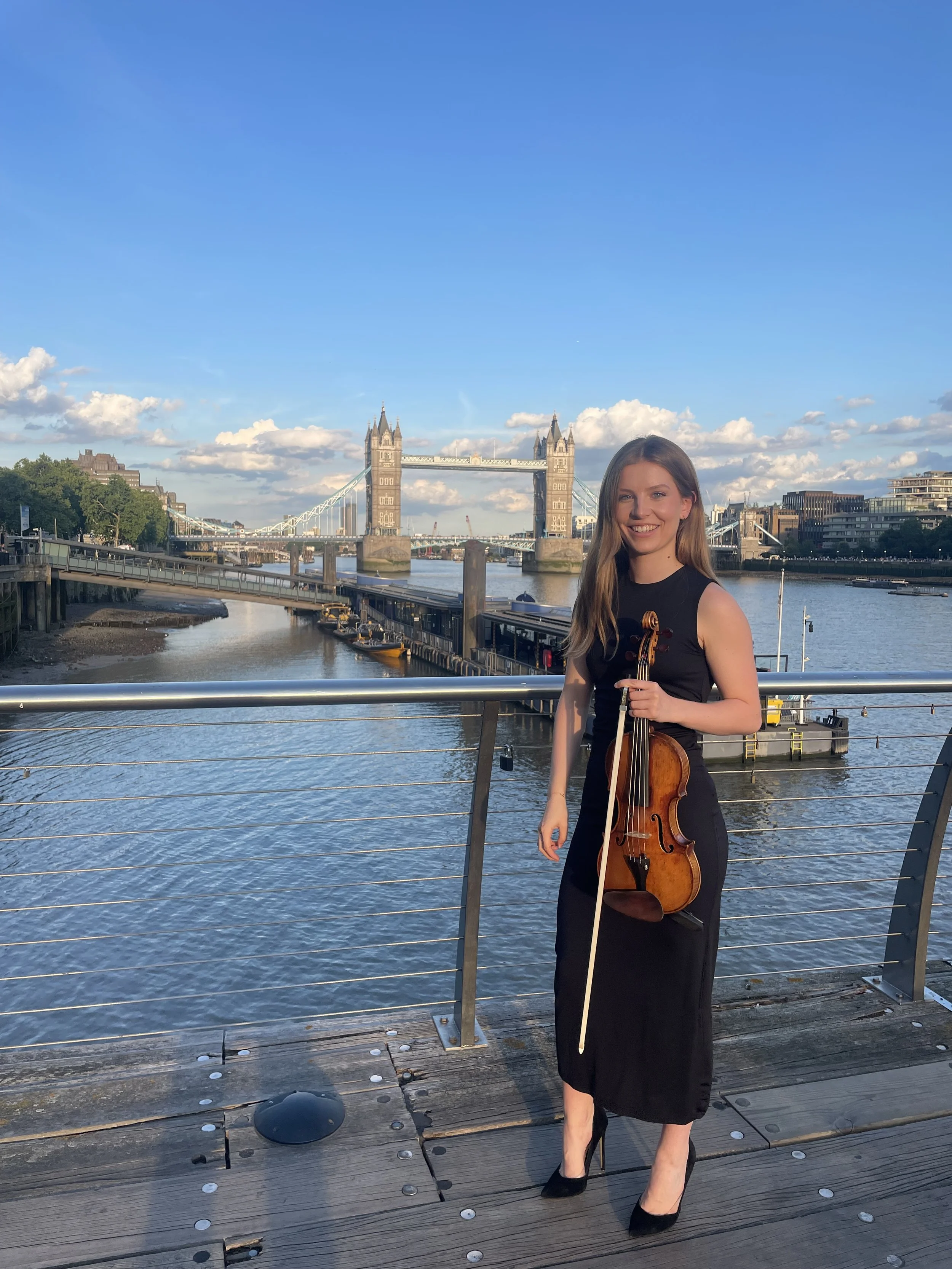 A woman in a black dress is standing on a wooden dock, holding a violin and bow, with Tower Bridge and the River Thames in London visible in the background.