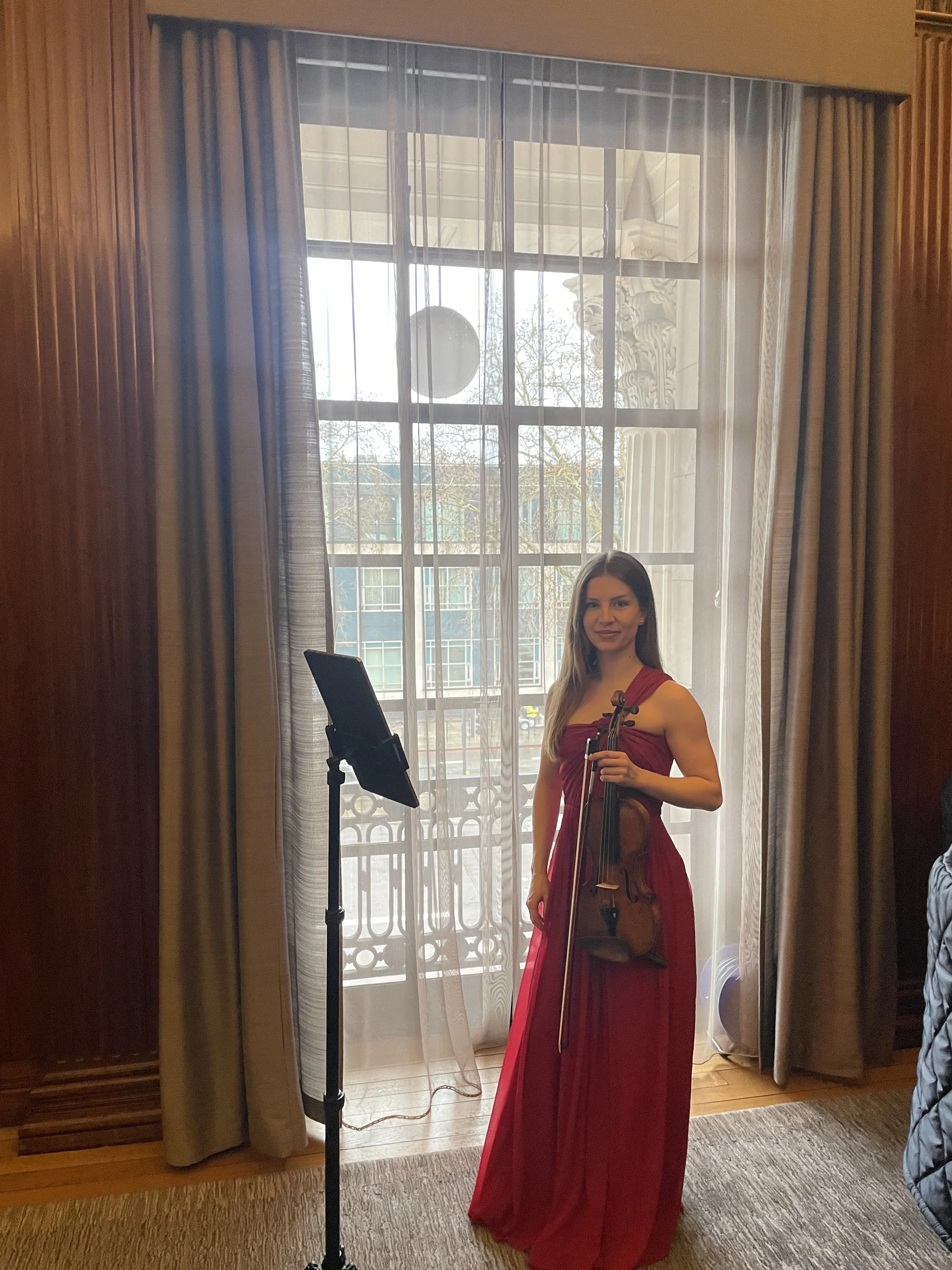 A woman in a red dress holding a violin, standing in front of a window with sheer curtains and a balcony outside, in an indoor setting.