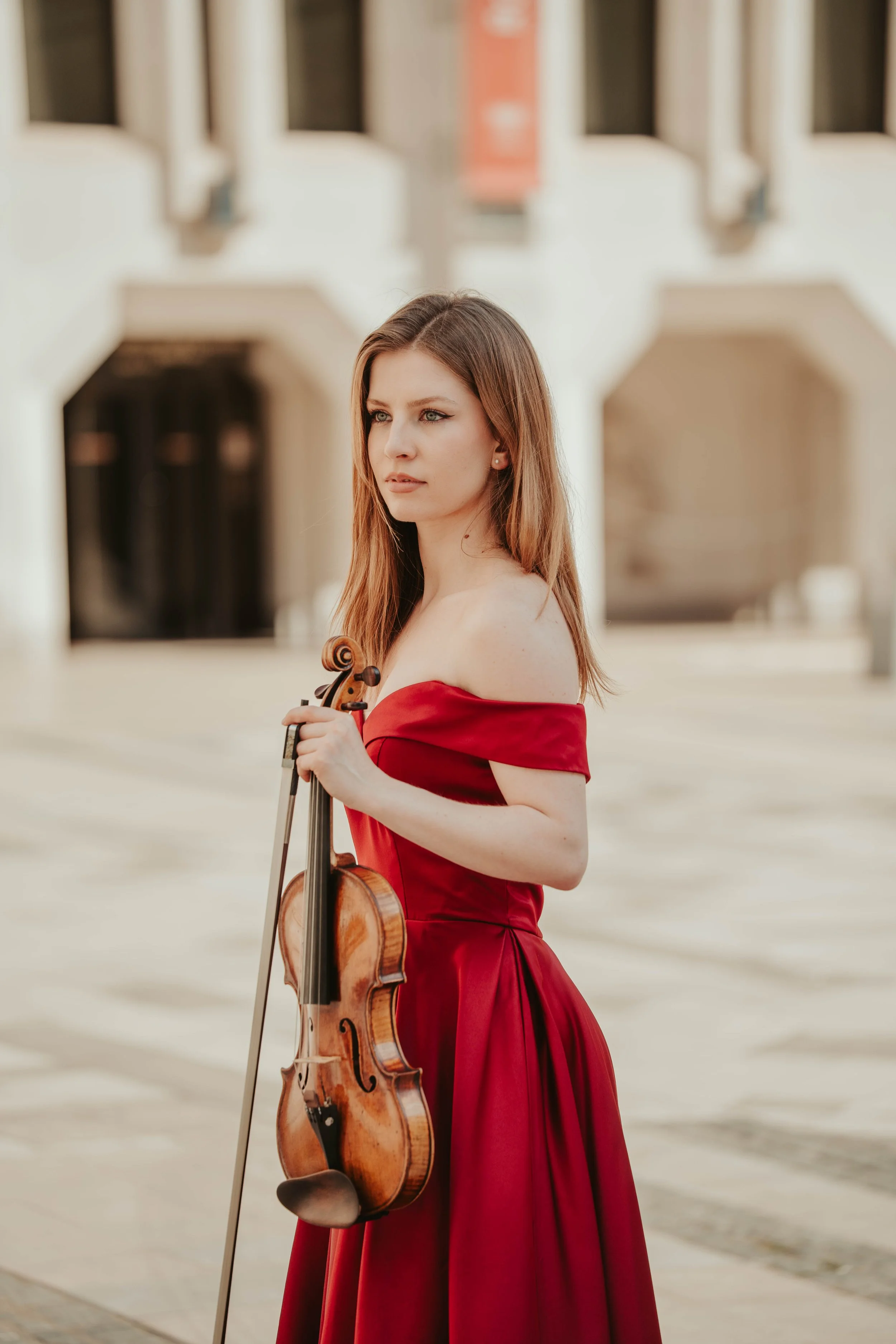 A young woman with long, light brown hair wearing a red off-the-shoulder gown holding a violin and bow outdoors in front of a classical building with arched windows.