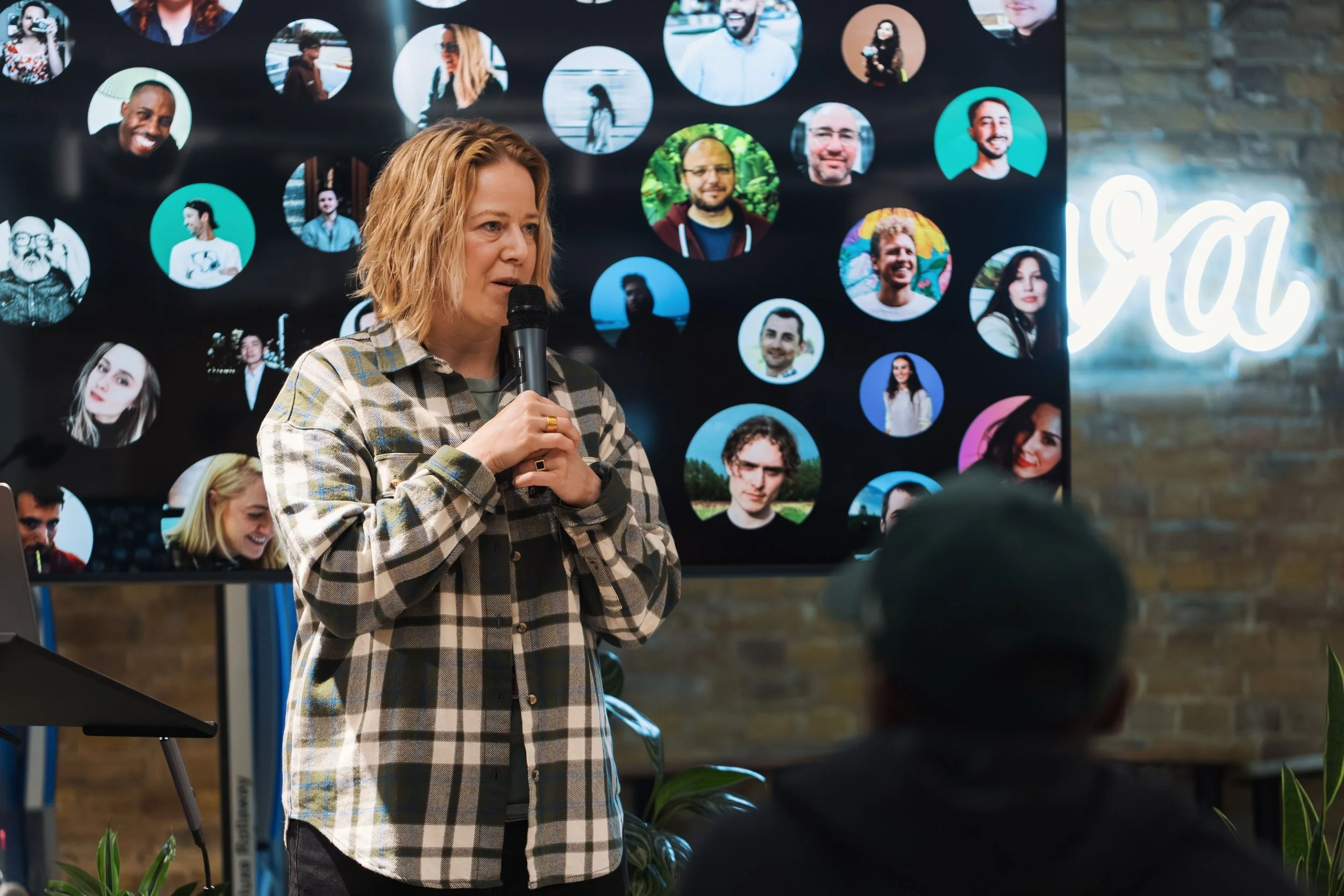 A woman with wavy blonde hair wearing a plaid shirt holding a microphone speaking at an event with a large screen behind her displaying various circular photos of diverse people. The background includes a neon sign with cursive writing and a brick wall.