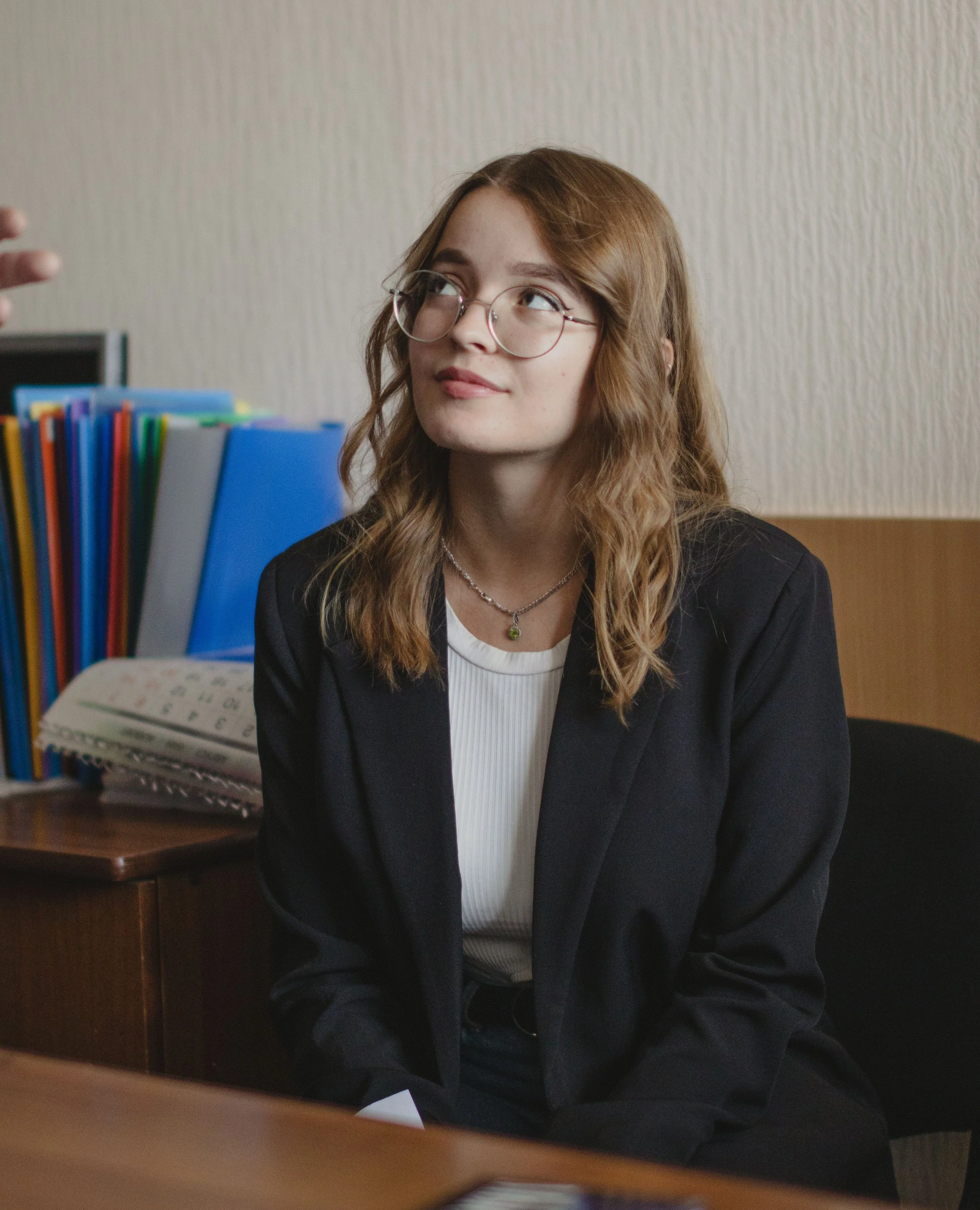 A young woman with wavy, reddish-brown hair and glasses, wearing a black blazer and a white top, sitting at a desk with a slight smile, in an office setting with light-colored textured walls and a bookshelf with colorful folders in the background.