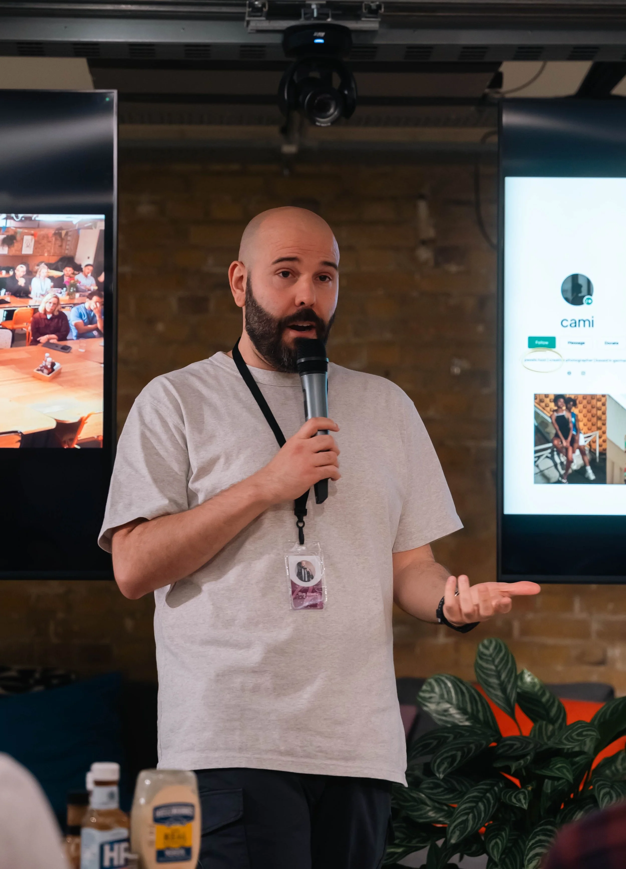 A man with a beard and a bald head is speaking into a microphone during a presentation or discussion at an indoor event. There are two large screens behind him displaying digital content.