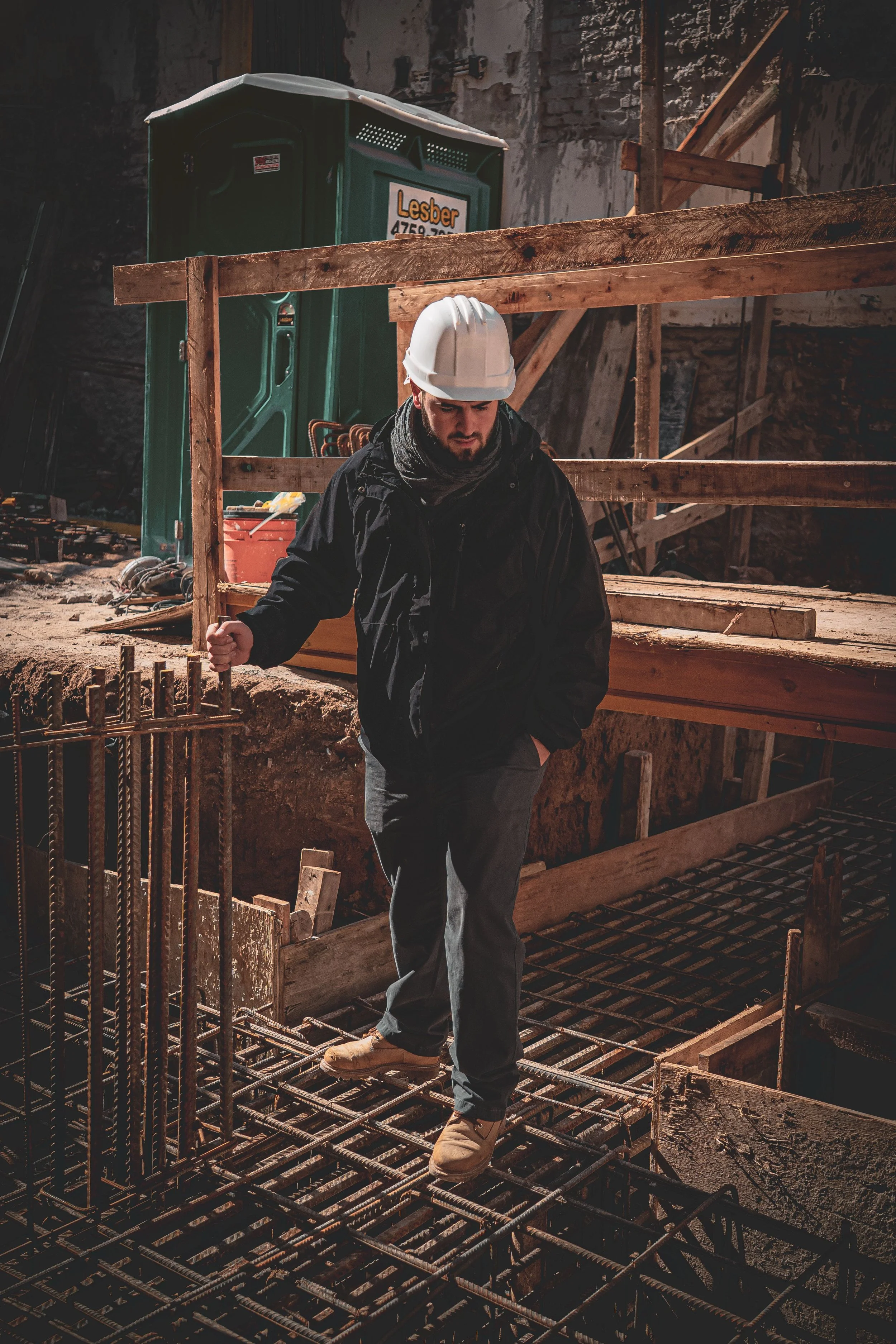 A construction worker wearing a white hard hat and black jacket standing on a rebar grid at a construction site with wooden framing and scaffolding in the background.