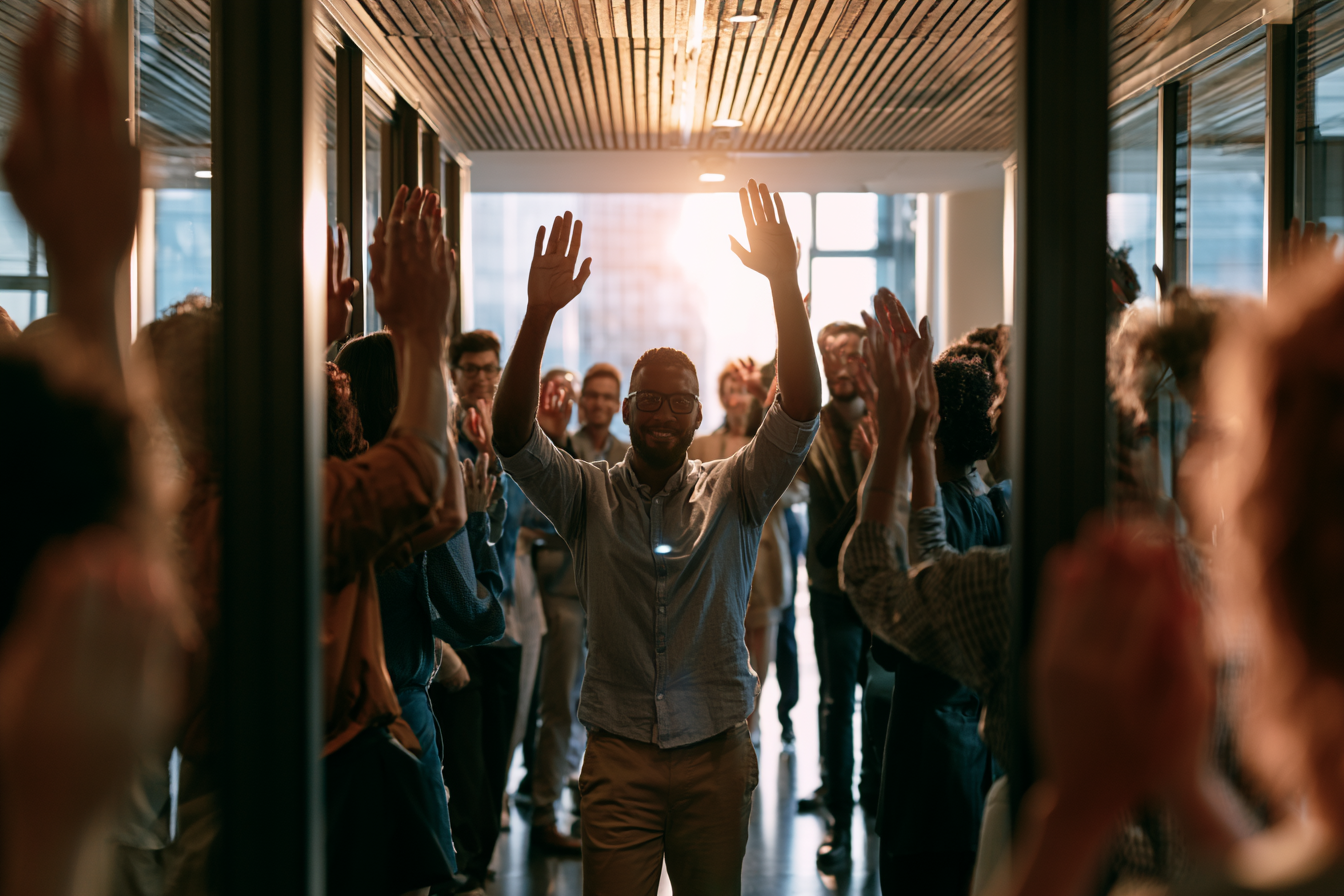 A diverse group of people celebrating and raising their hands in a modern office space during daylight.