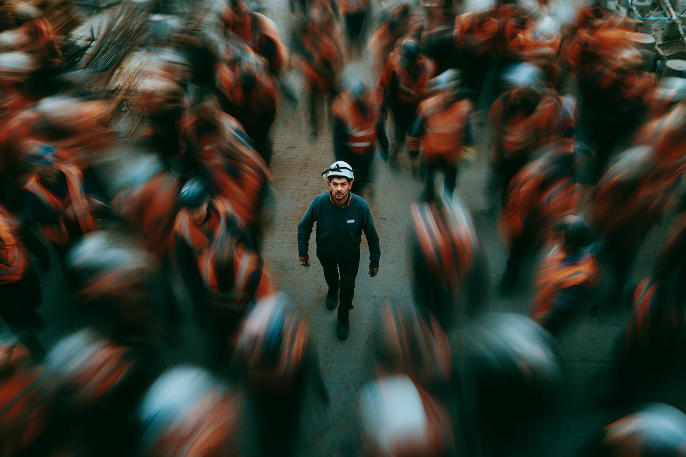 A man wearing a helmet and dark clothing walking through a crowd of workers dressed in orange uniforms, captured from above with motion blur.