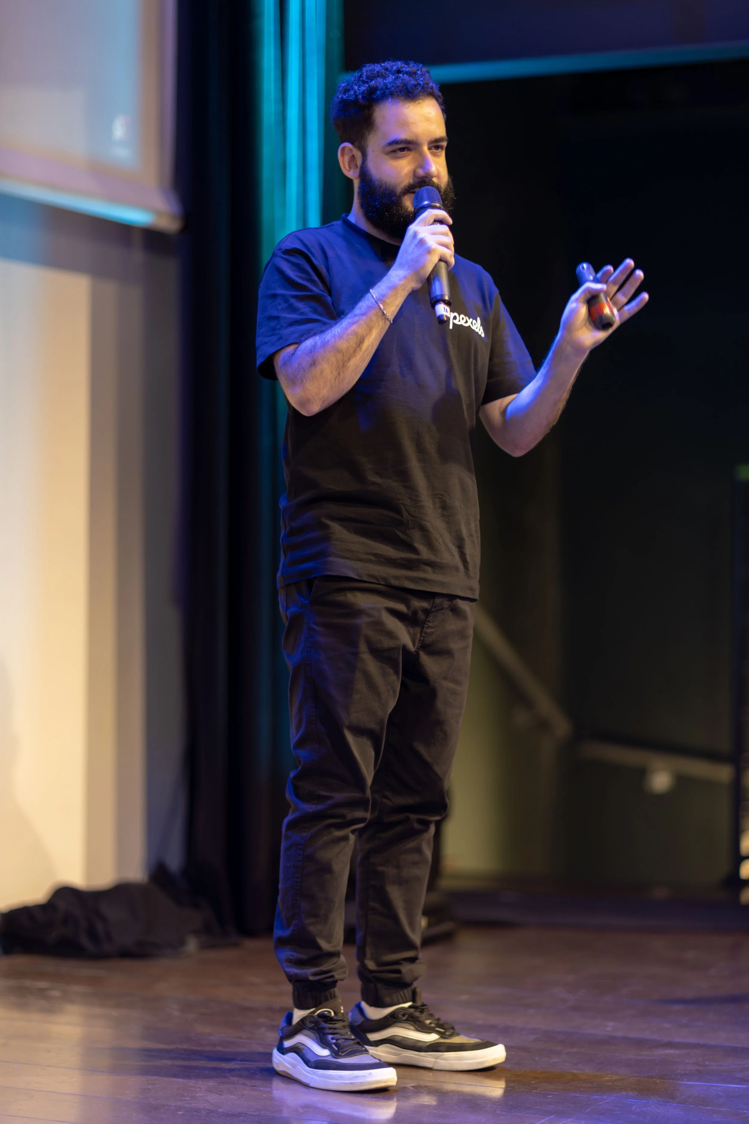 A man with a beard, wearing a black t-shirt and black pants, is standing on a stage holding a microphone and gesturing with his left hand, with a projector screen and dark curtains in the background.