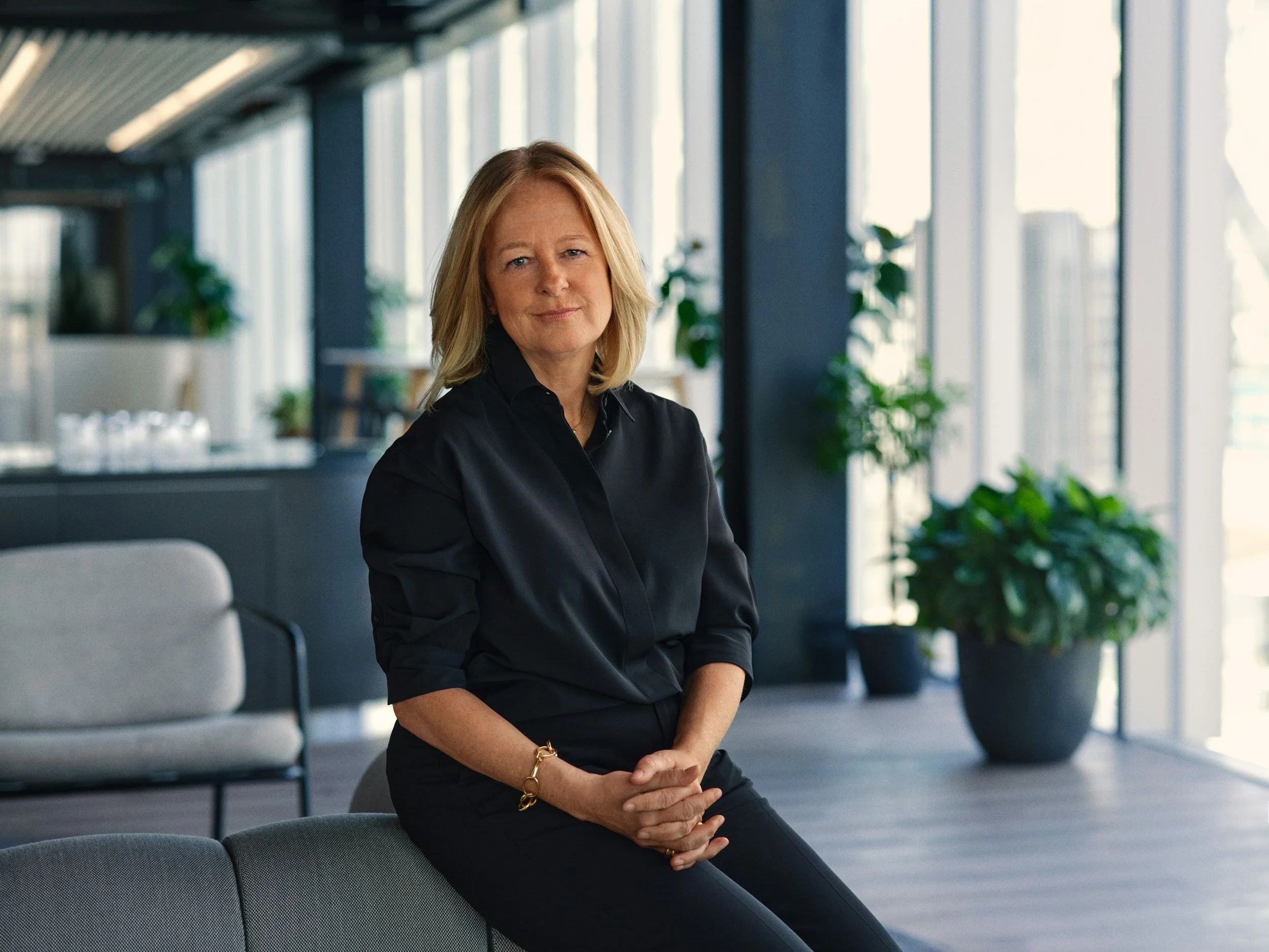 A woman with blond hair sitting in a modern office, wearing a black shirt, with a neutral expression, in front of large windows with natural light and green plants in the background.