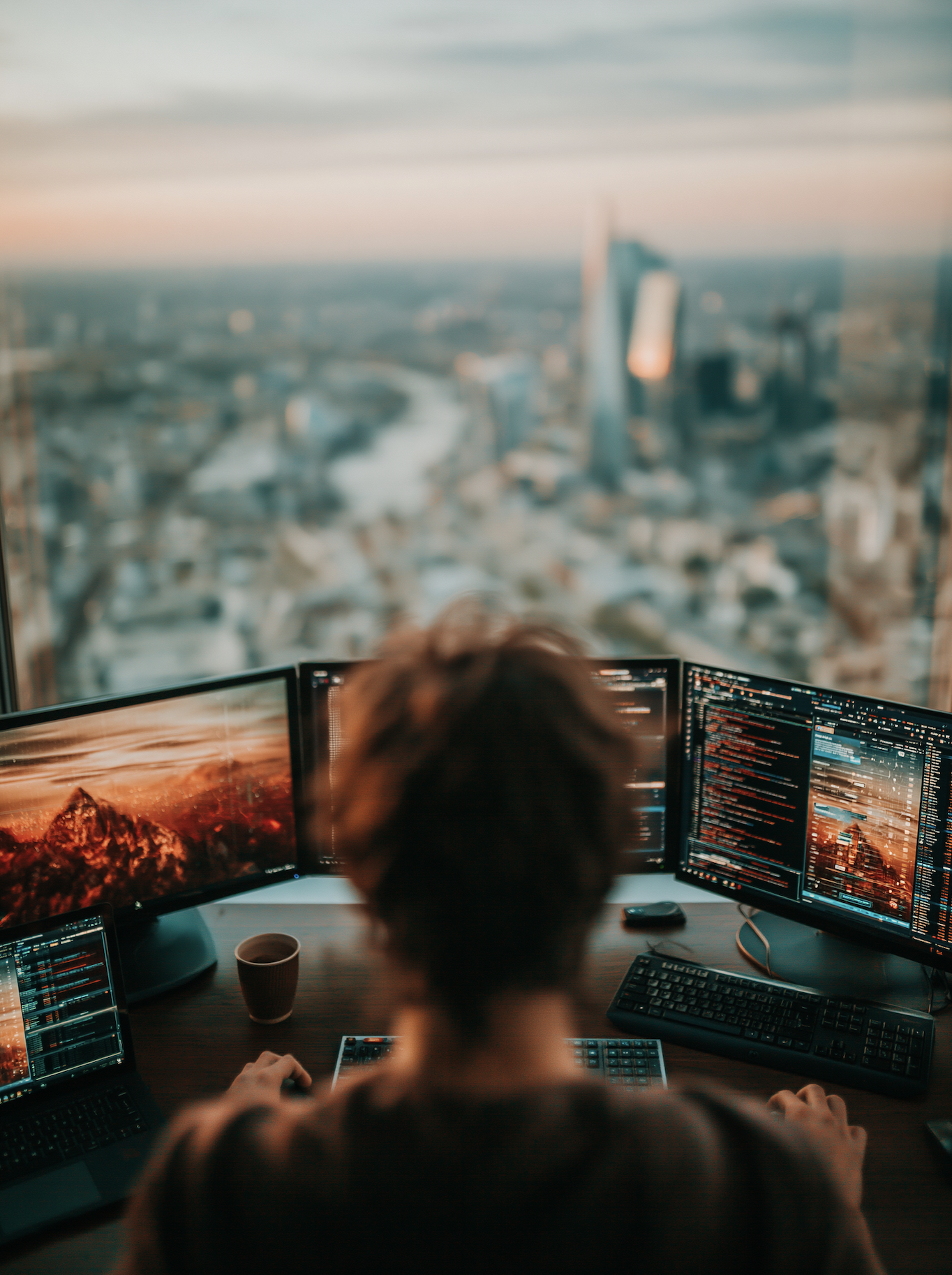 A person working at a desk with three monitors displaying code, with a city view and skyscrapers visible through a large window in the background.