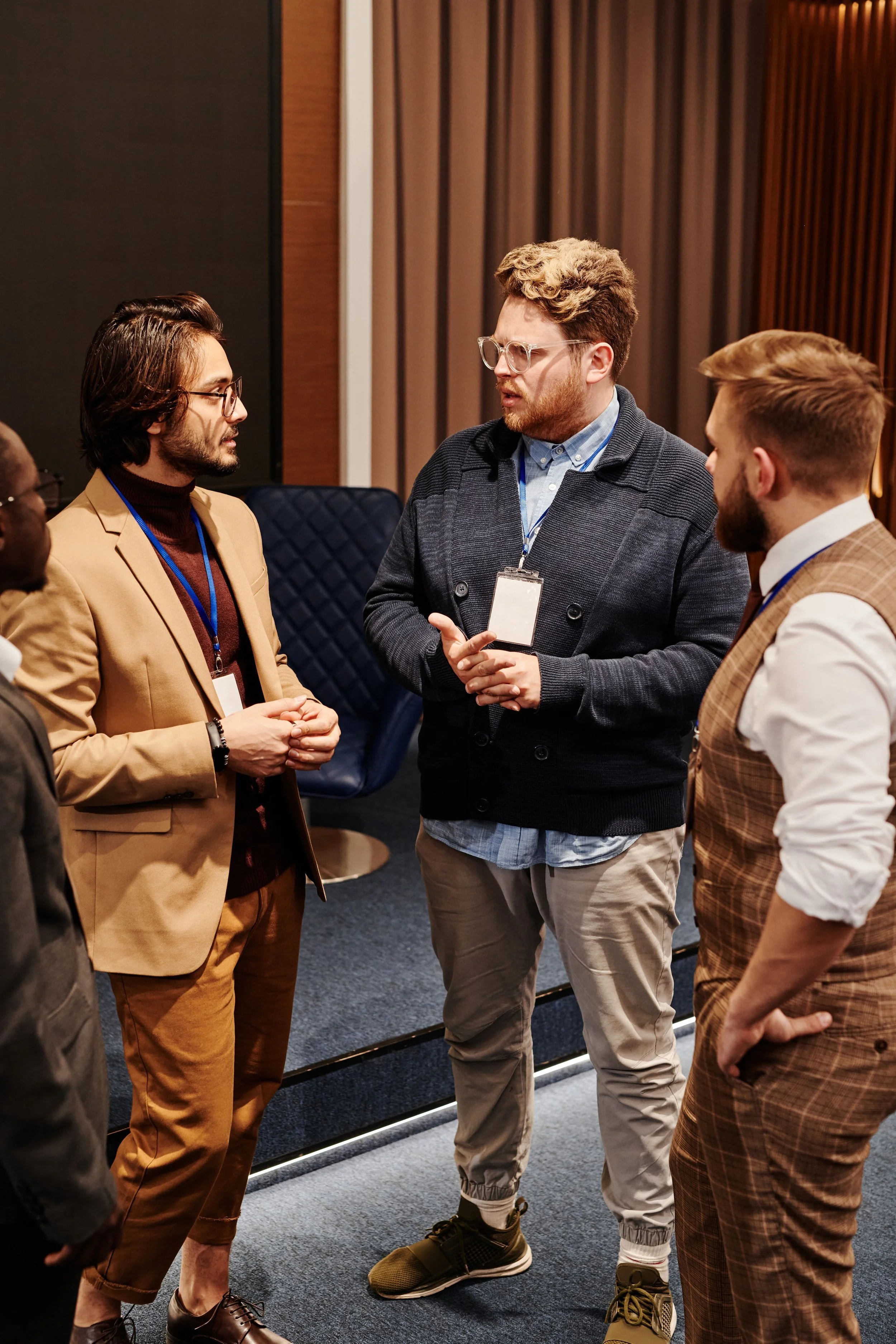 A group of five diverse men in business casual attire, standing in a circle and engaging in conversation in a professional indoor setting with dark carpet and curtains.