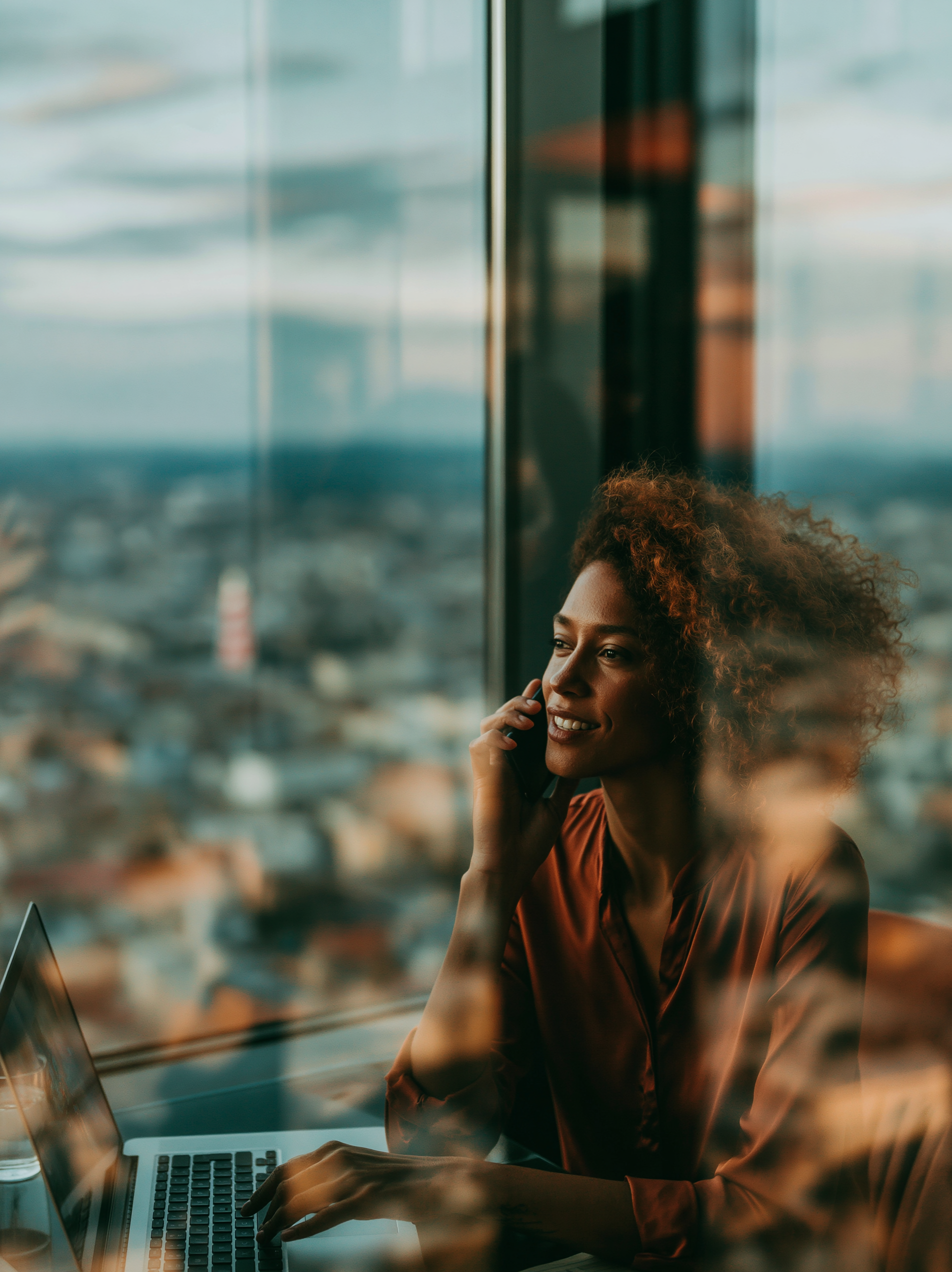 A woman with curly hair talking on her cellphone while working on a laptop at a desk near a large window with a city view.