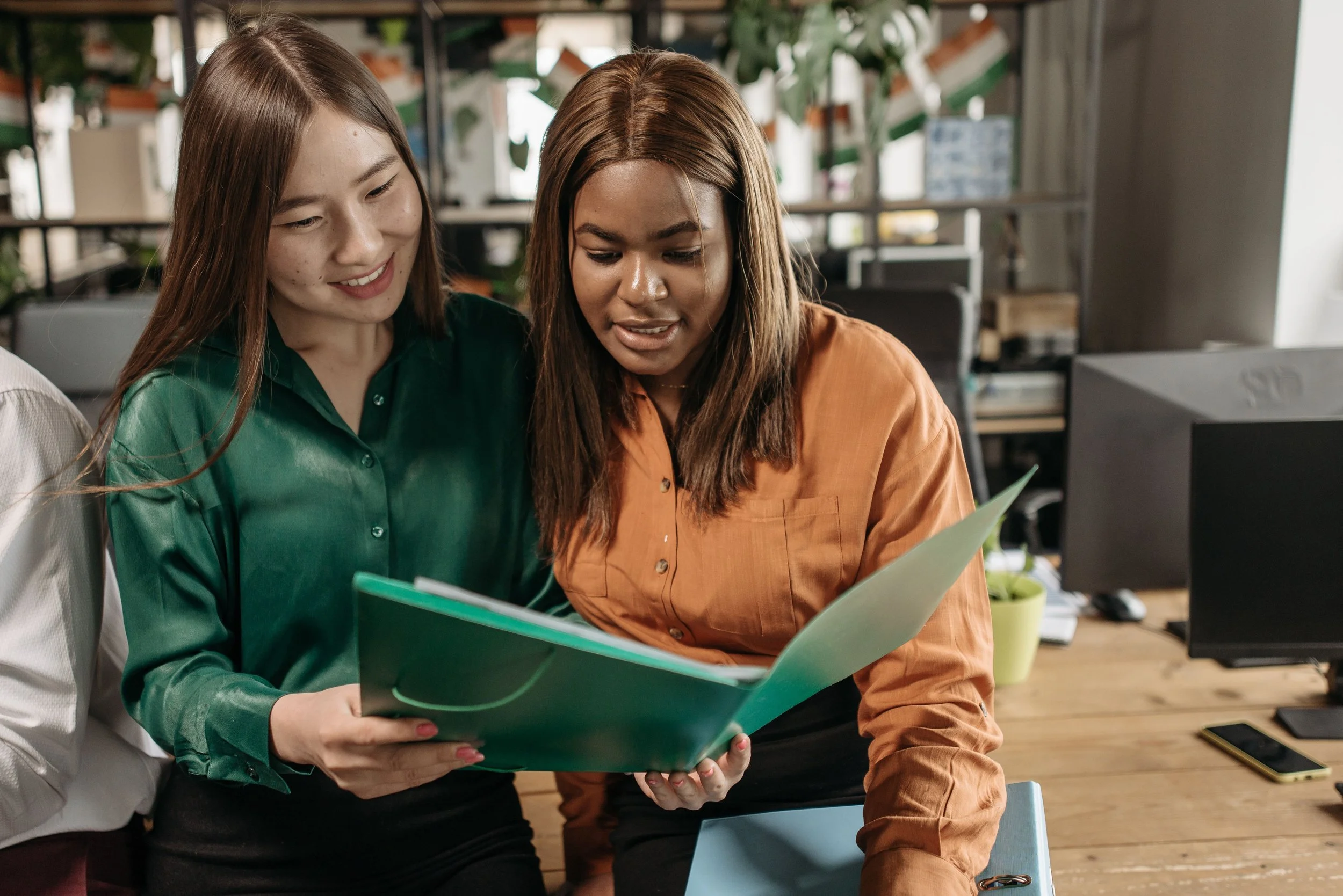 Two women sitting together in an office, looking at a green folder or book.