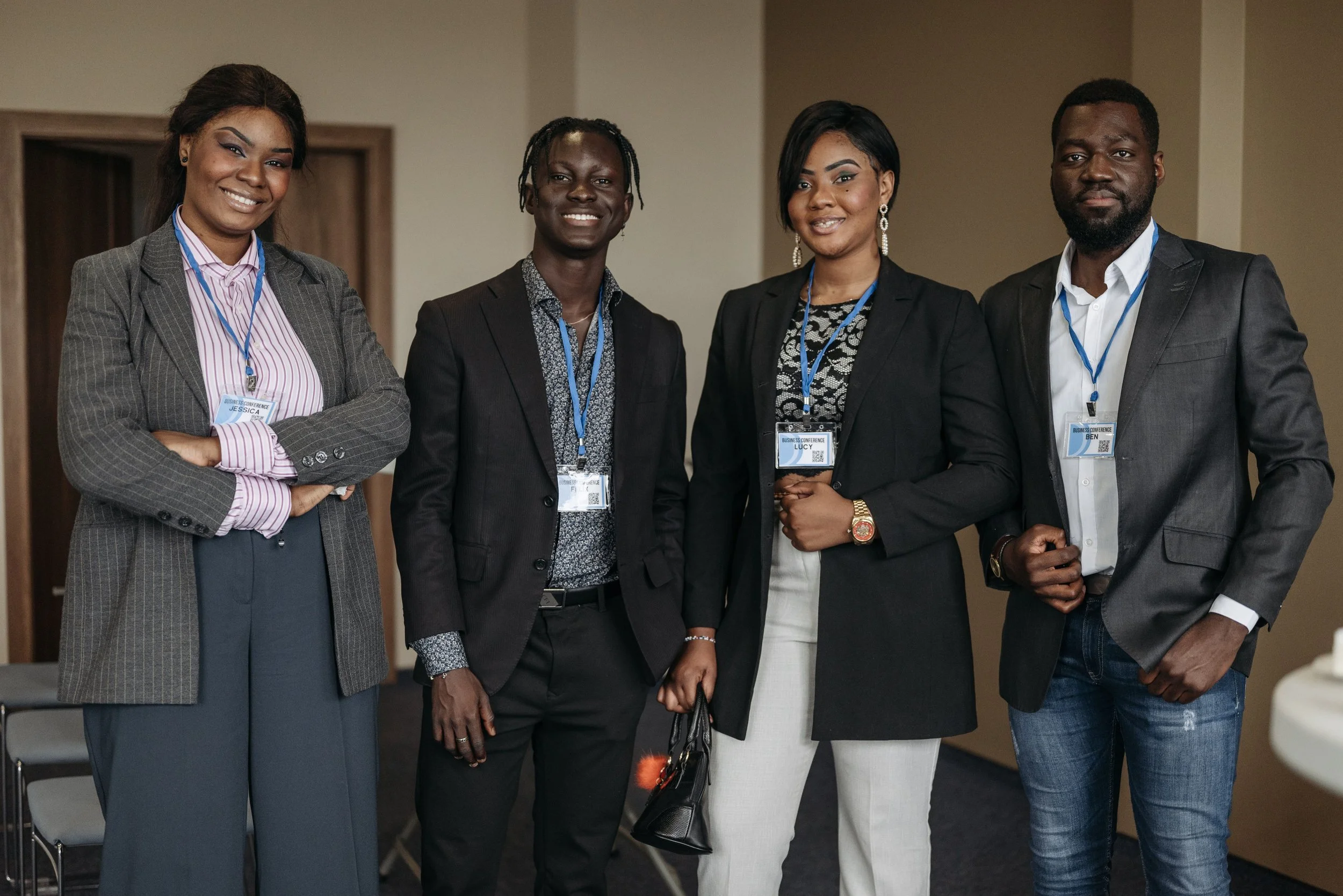Group of four diverse professionally dressed individuals at a conference, wearing name tags, standing in a line inside a conference room.