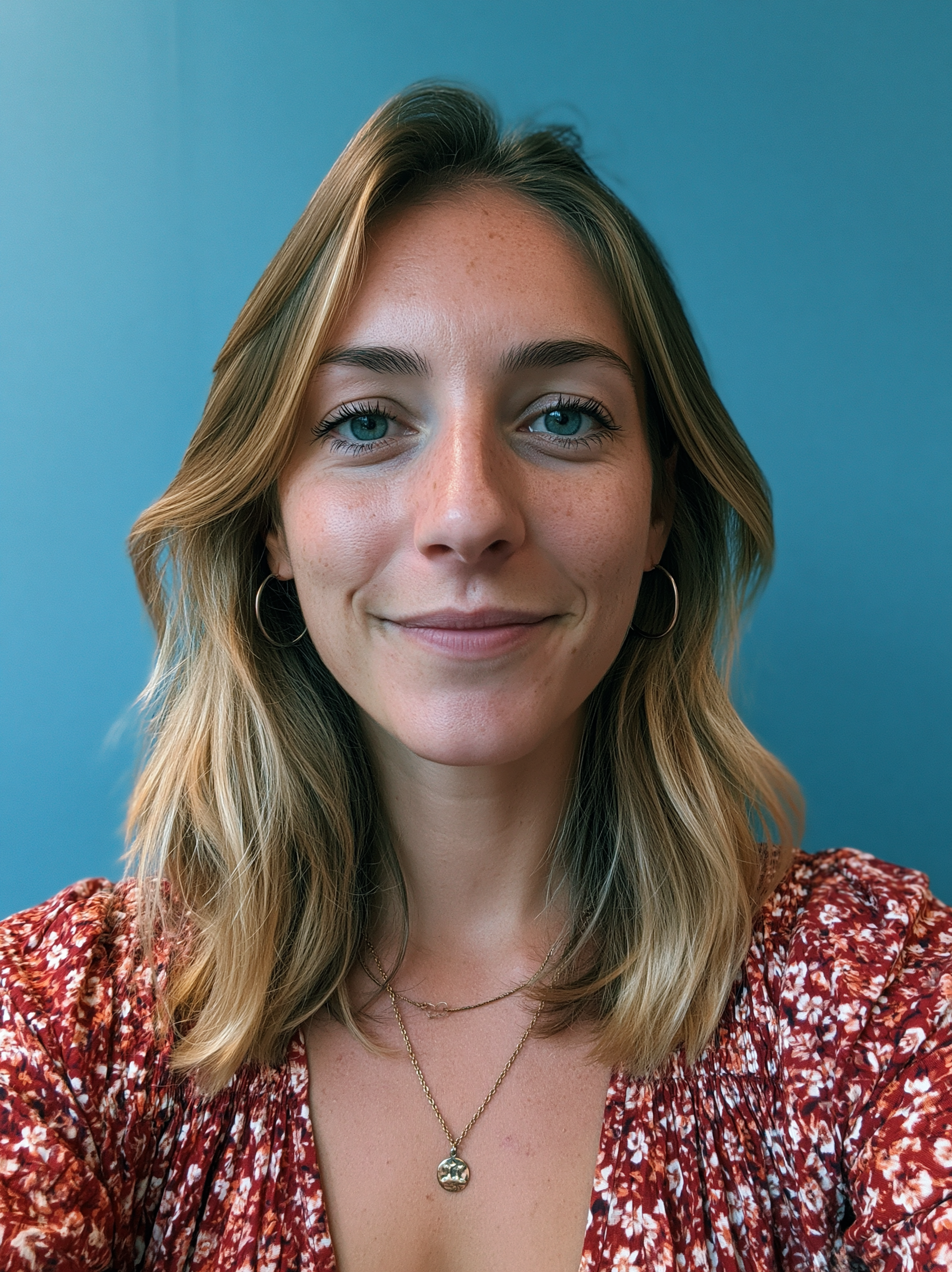 Close-up of a young woman with blonde hair and blue eyes, wearing a red patterned top and layered gold necklaces, smiling softly against a blue background.