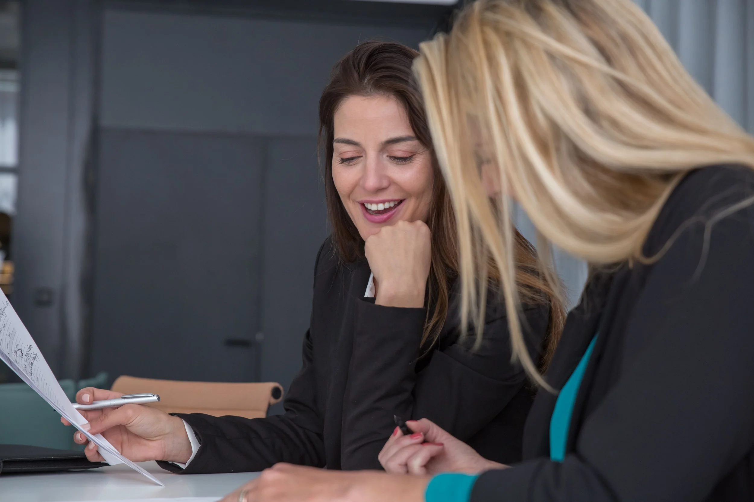 Two women are talking and smiling while looking at papers on a table in an office setting.