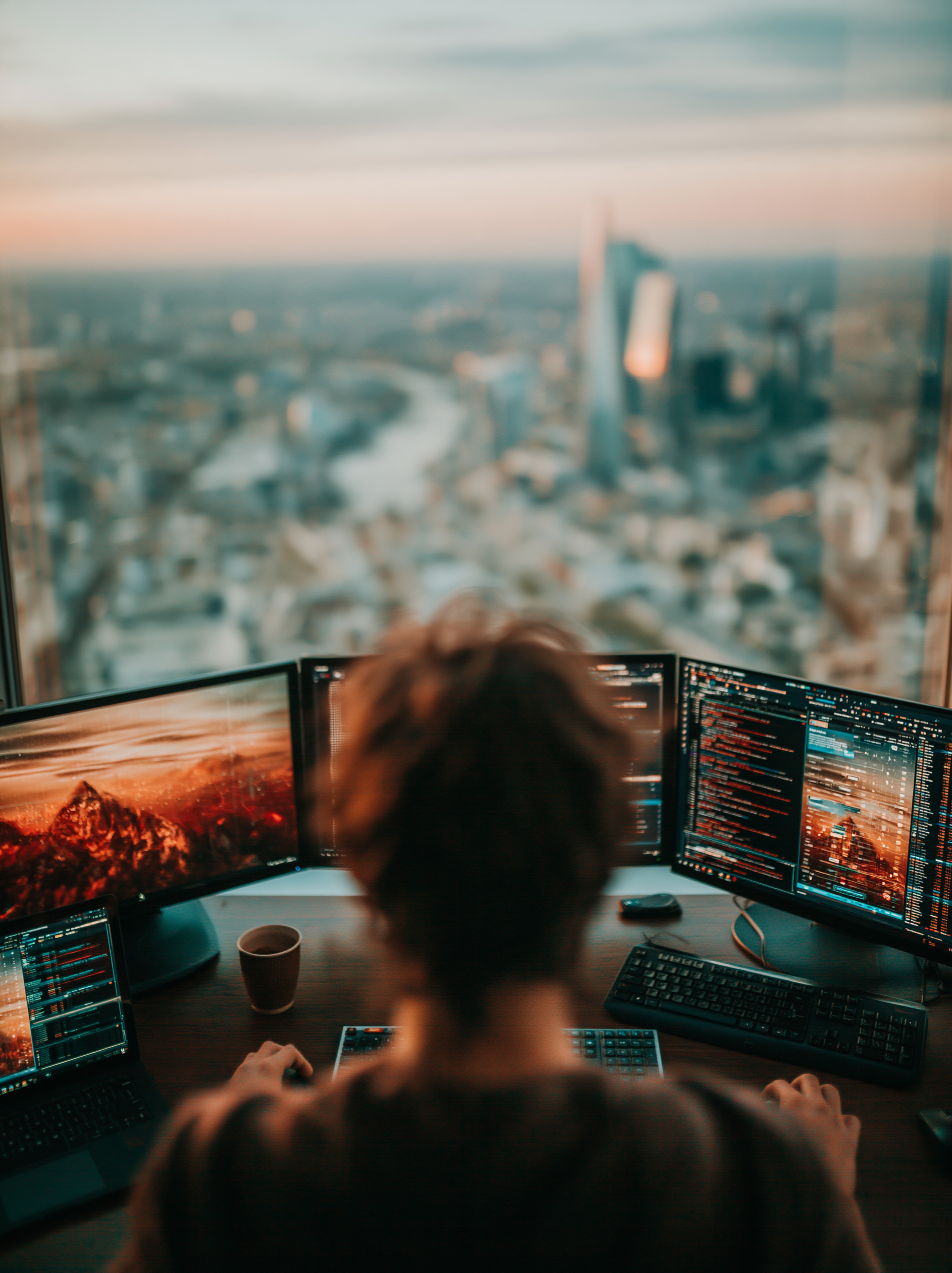 Person working at a desk with multiple computer monitors displaying code, in an office with a city skyline view through a large window.