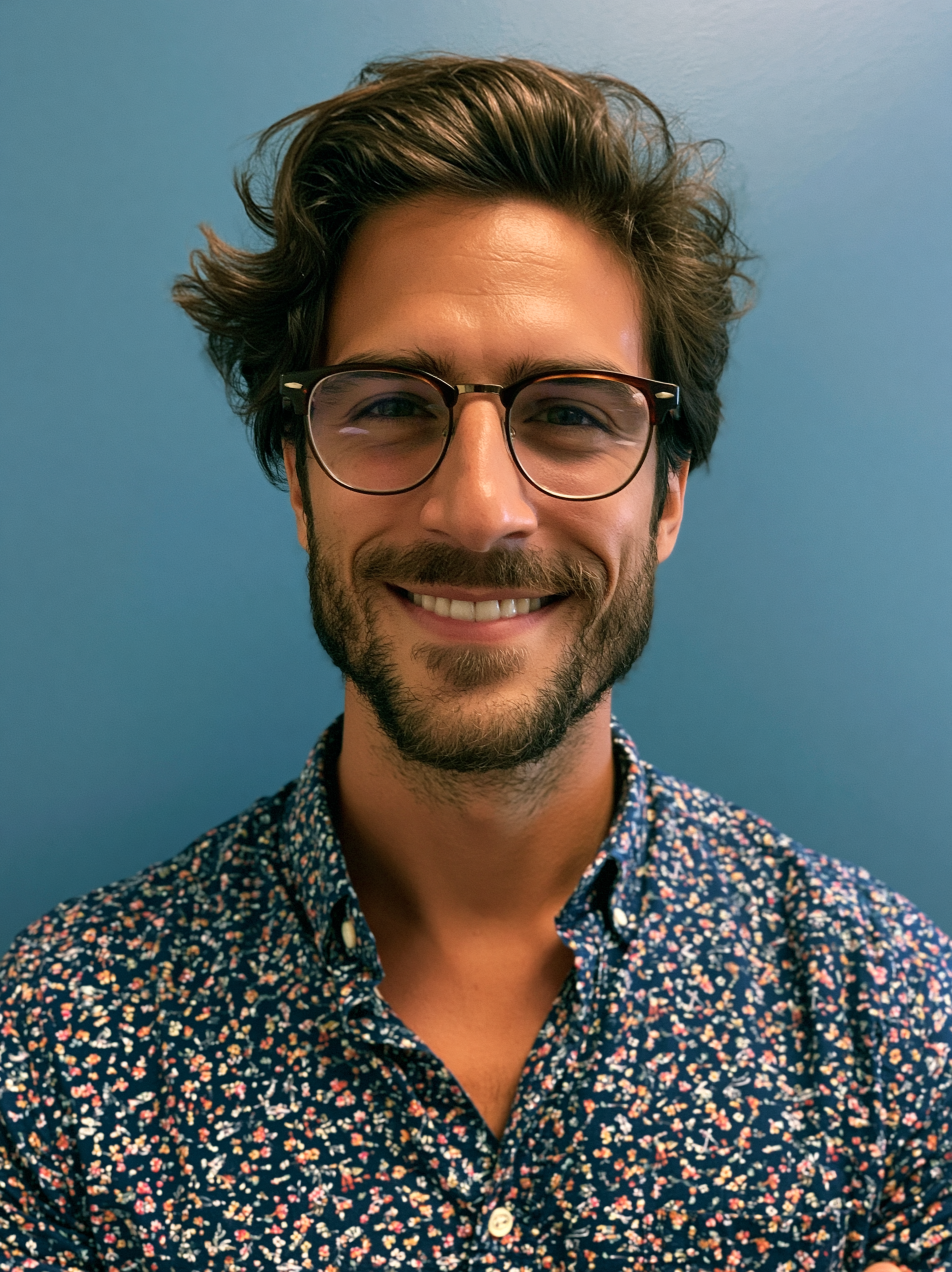 A man with light skin, wearing glasses with a dark frame, smiling, with dark brown wavy hair, a beard, and a colorful floral shirt, standing against a light blue background.