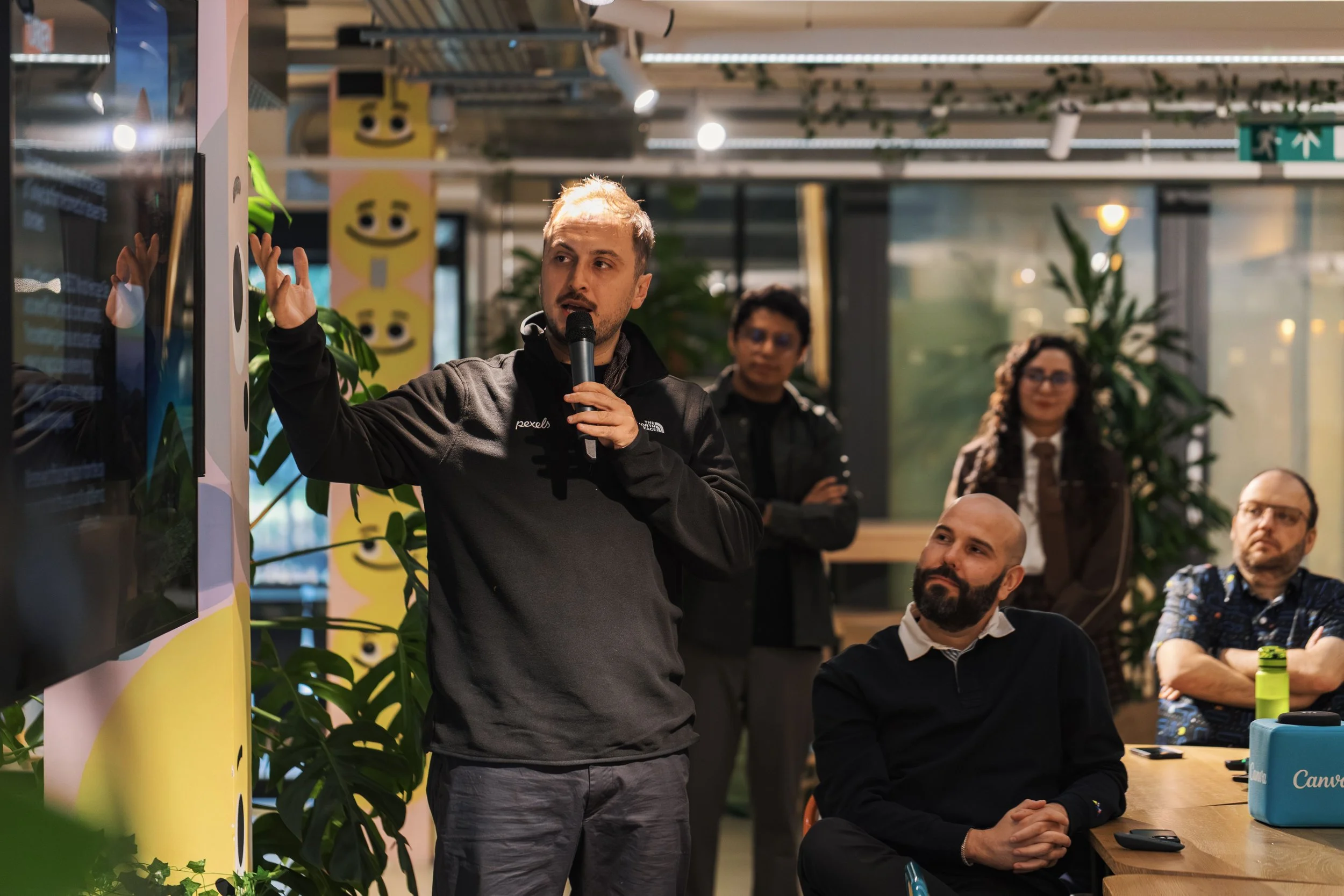 A man in a black jacket is speaking into a microphone while gesturing with his hand during a presentation. Several people are seated around the table, listening attentively in an indoor setting decorated with green plants and colorful artwork.