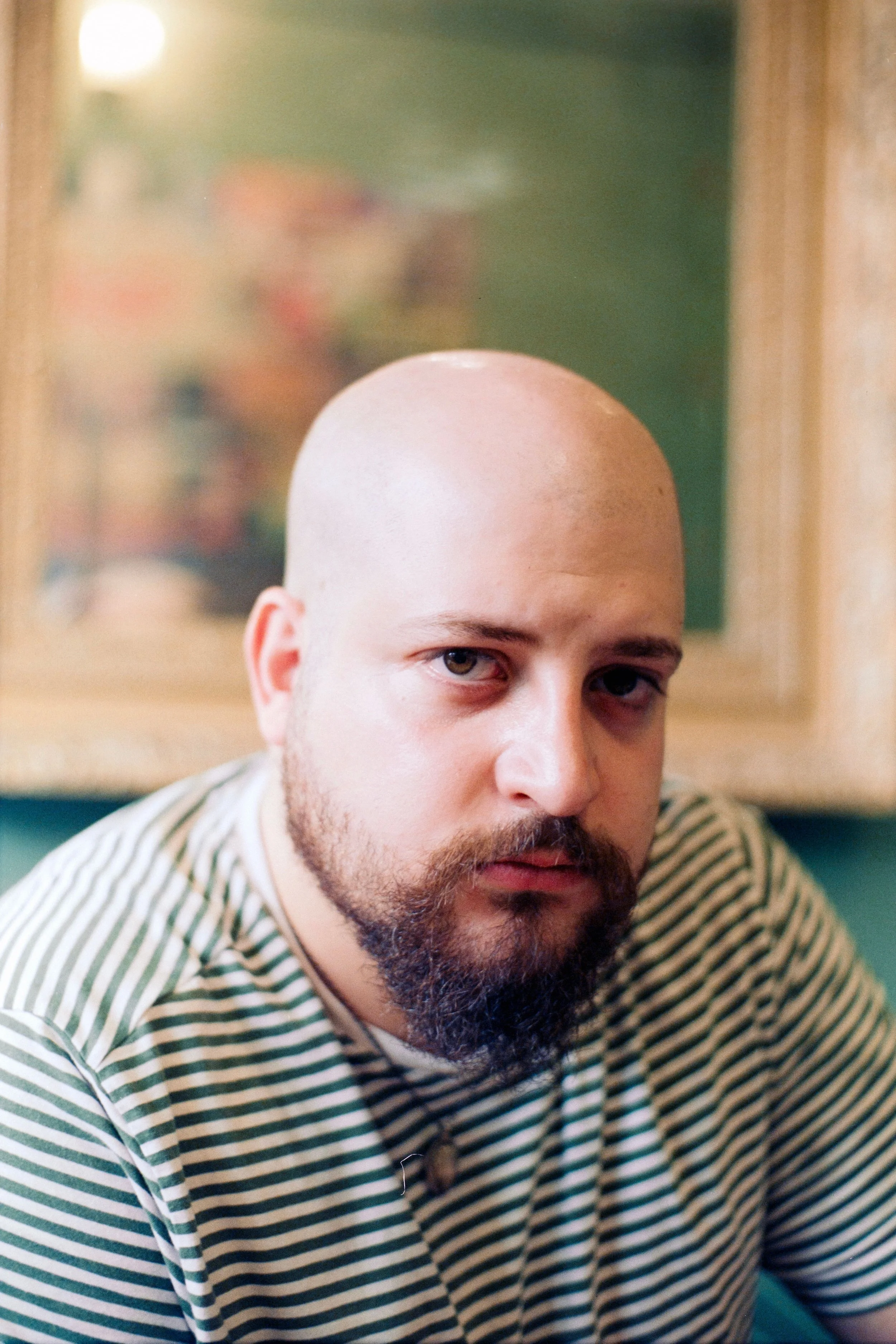 A man with a beard and no hair, wearing a striped shirt, is looking into a mirror, with the top half of his face and head visible. The background is blurred with green and wooden tones.