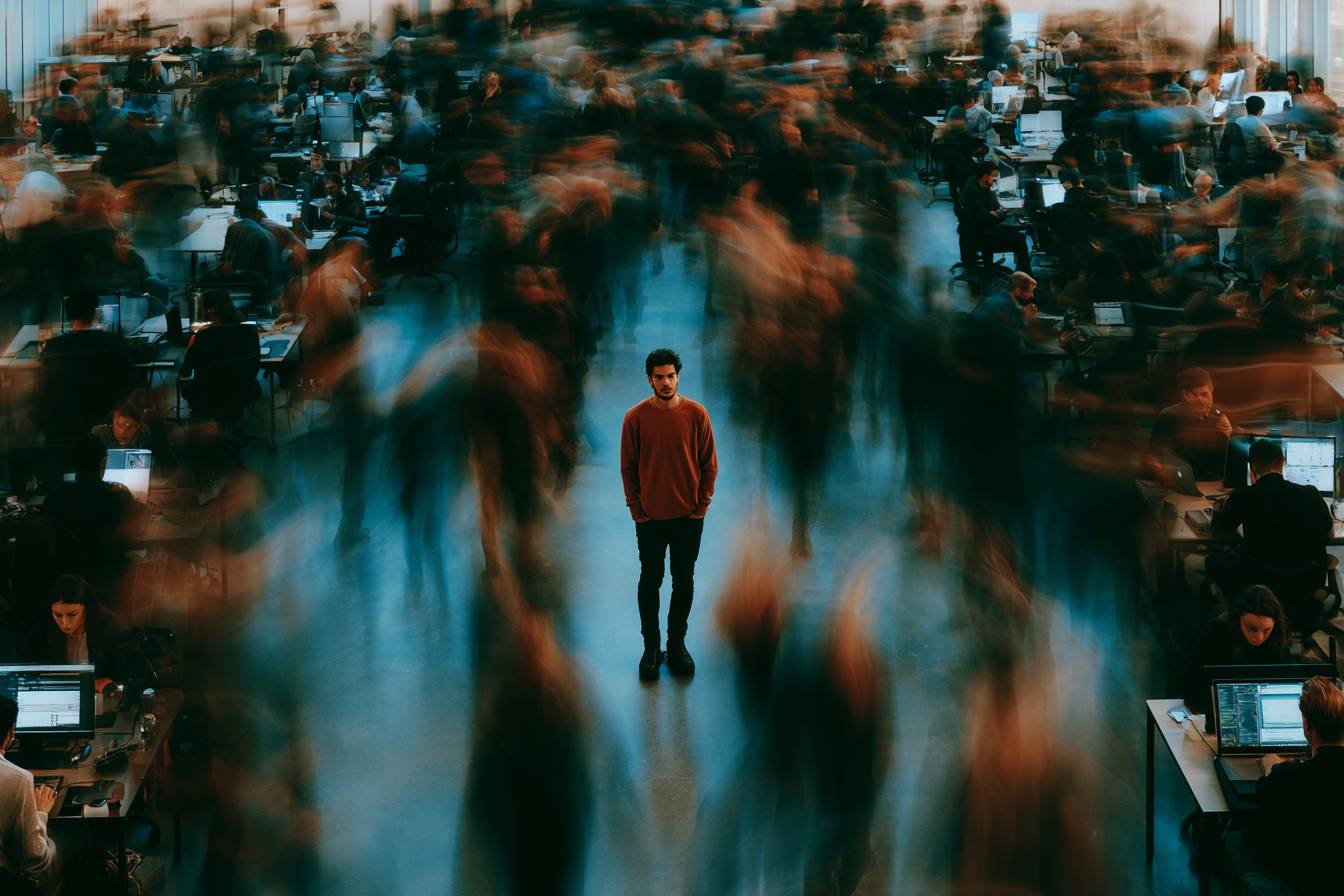 A man in a red sweater standing alone in the center of a busy, crowded office space with many people working on computers, some blurred to indicate movement.