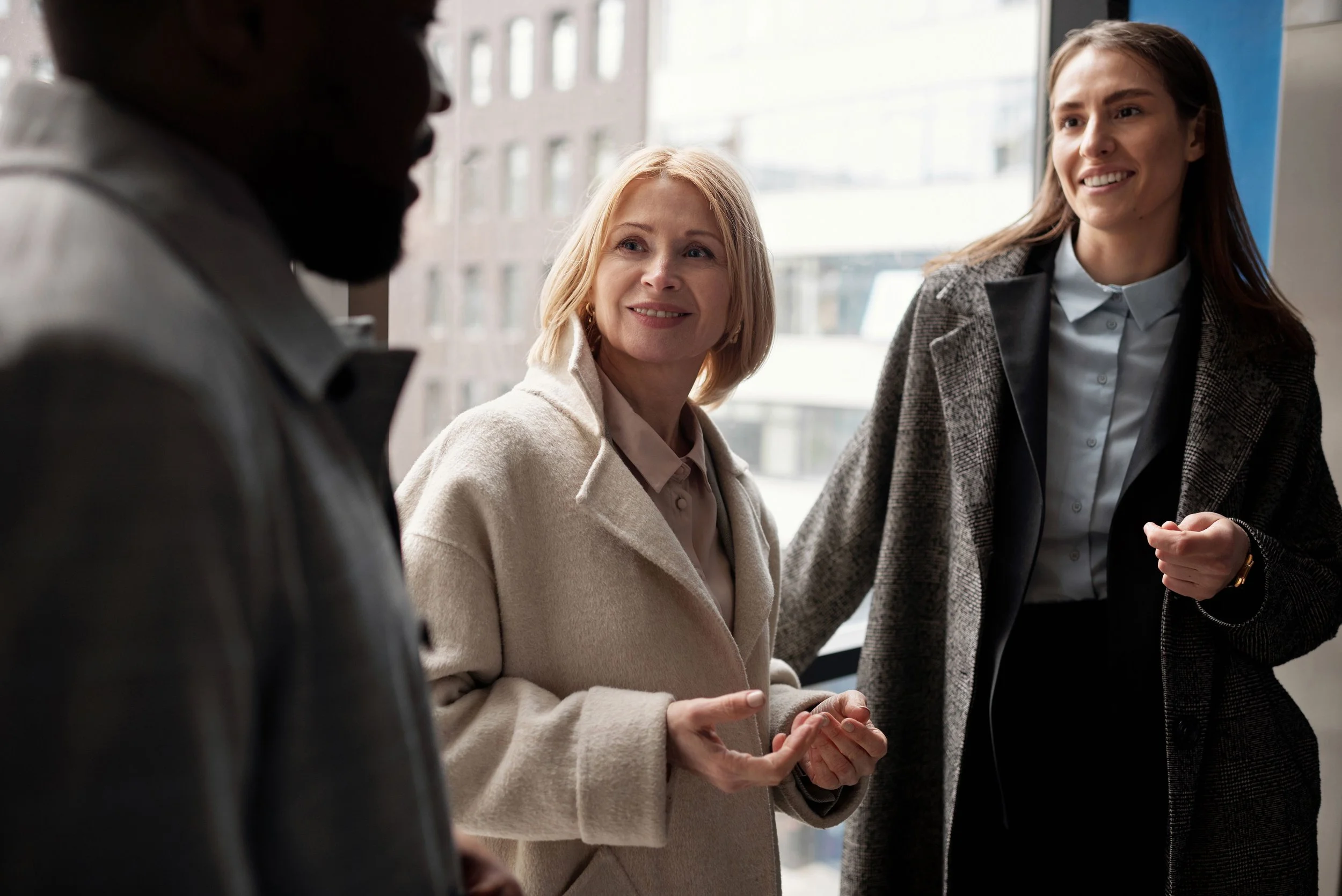 Three women, one blonde and two with brown hair, are having a conversation in an office with large windows in the background.