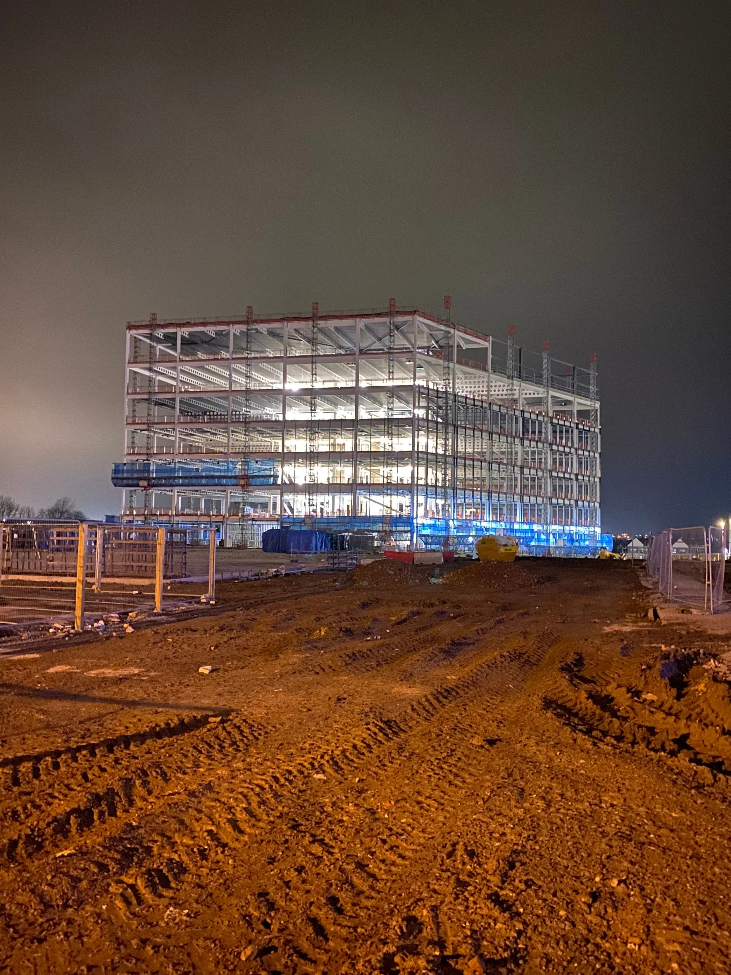 Nighttime construction site with a partially built multi-story building illuminated from inside, with dirt ground and construction barriers in the foreground.
