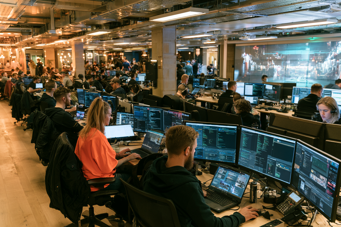 A busy office filled with people working on multiple computer monitors, with a large screen showing a cityscape in the background