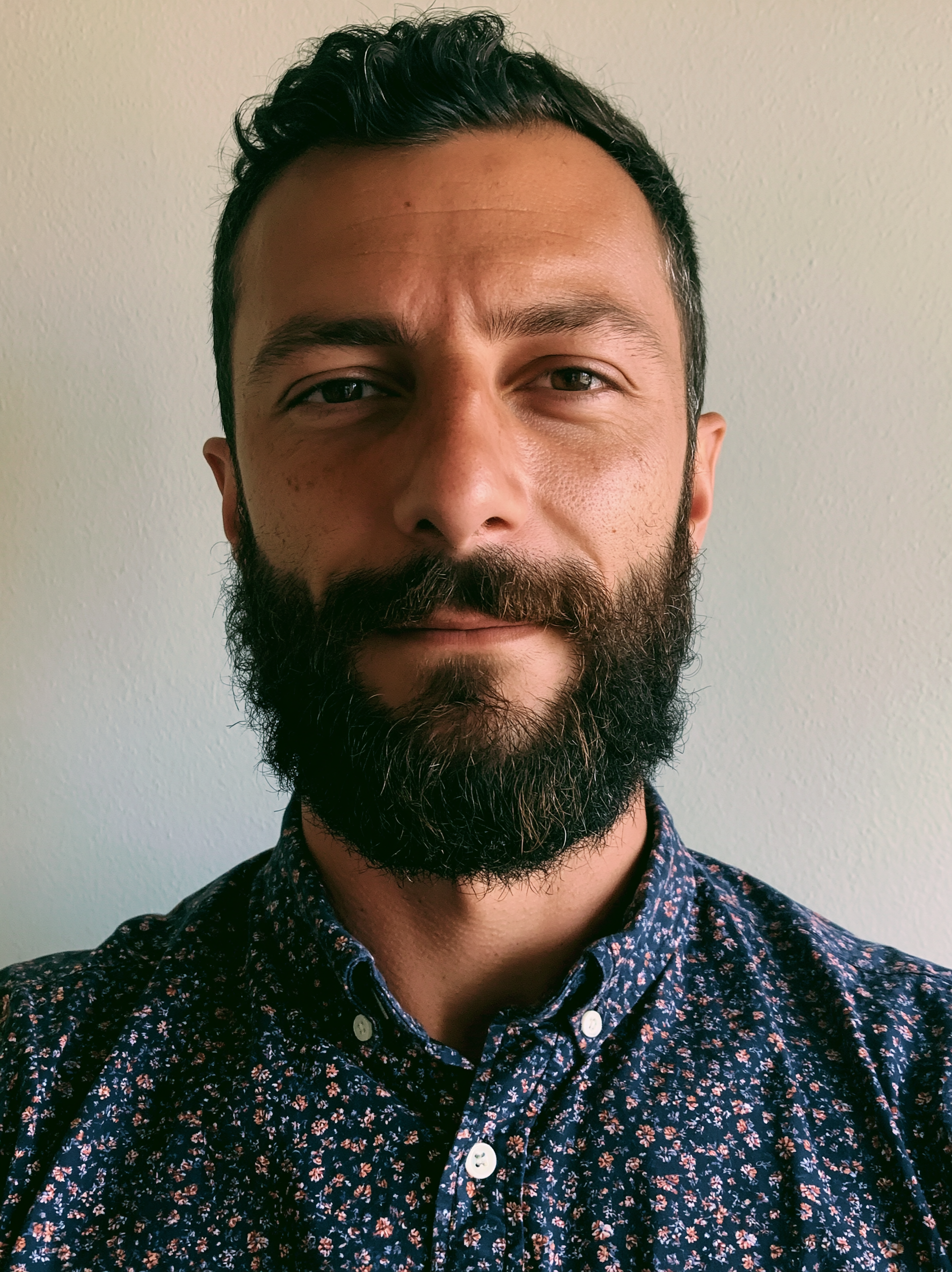 Close-up photo of a man with dark, wavy hair, brown eyes, and a full beard, wearing a dark floral patterned shirt against a plain light-colored wall.