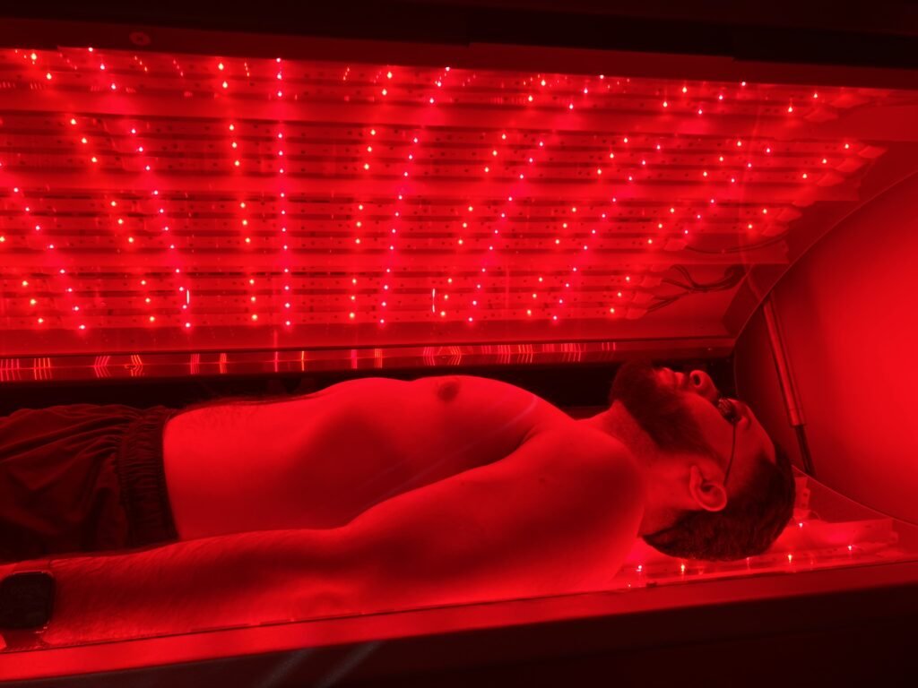 A shirtless man lying inside a red LED light therapy chamber experiencing Live Superhuman Experience.