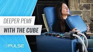 Woman reclining in a medical chair with a pillow, undergoing a Pulse XL Pro PEMF DEEPER PEMF therapy session with a device labeled 'WITH THE CUBE'