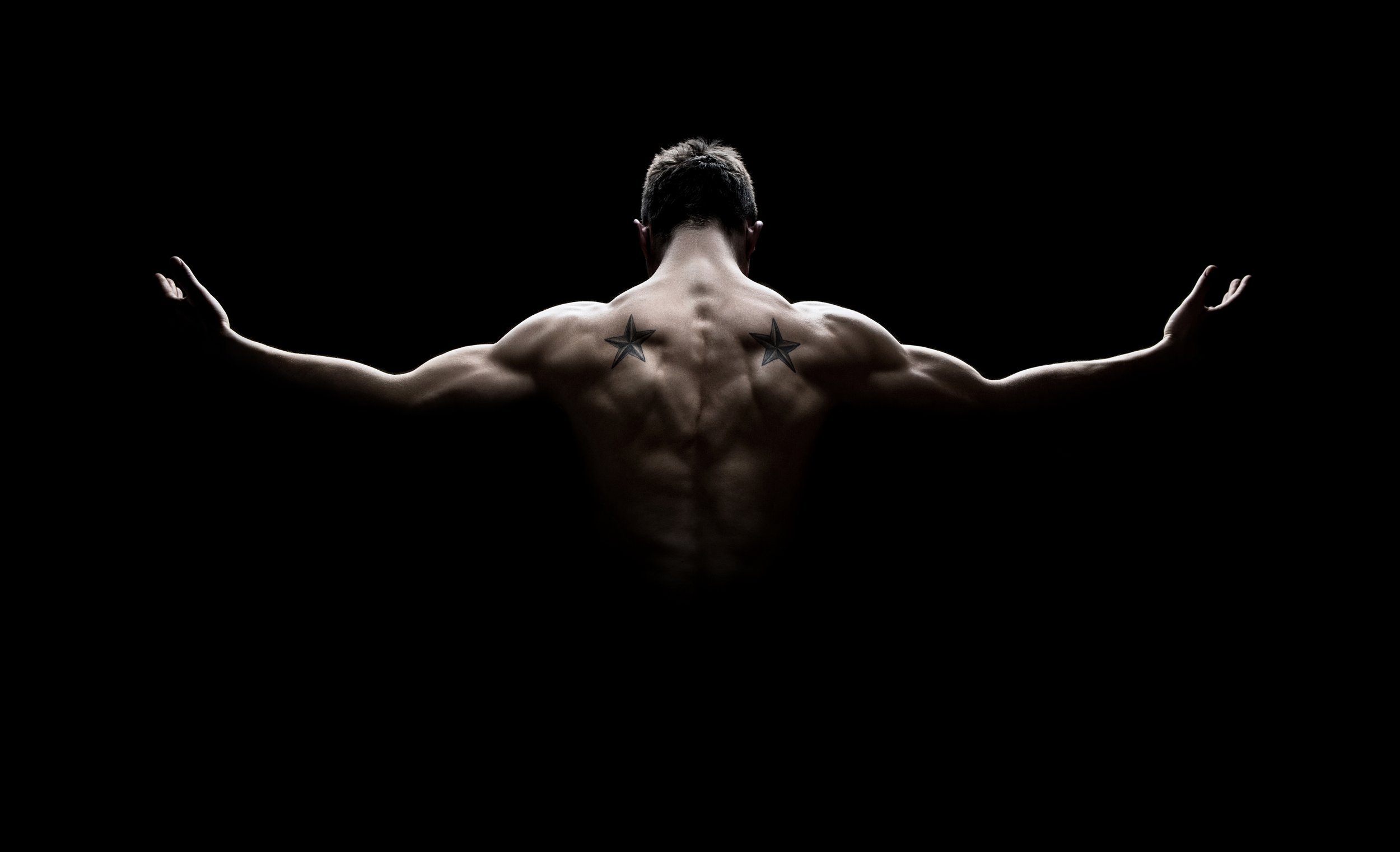 Back view of a man with arms outstretched representing LIVE Superhuman Experience against a black background, showing muscular physique and two star tattoos on shoulders.