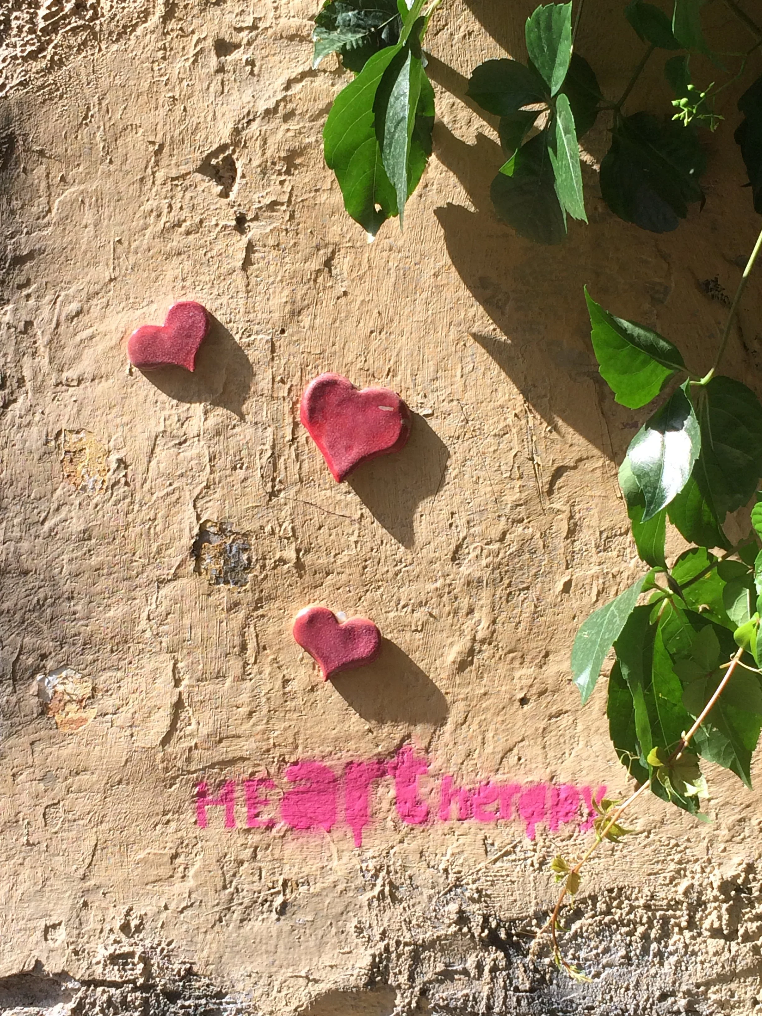Grüne Blätter und rote Herzen an einer alten Steinmauer, darüber pinke Sprühfarbe und dem Wort HEartherapy