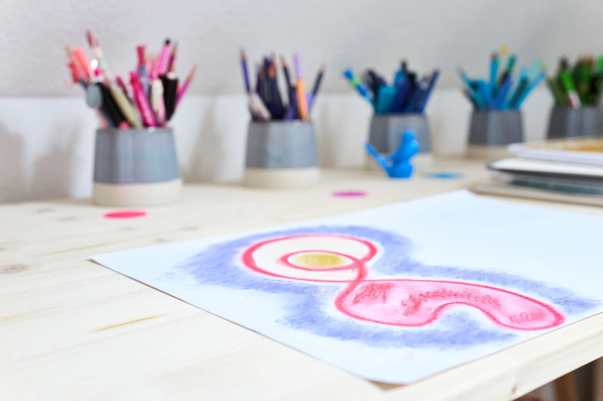 Bunte Filzstifte in grauen Töpfchen und eine Zeichnung mit farbigen Kreisen auf einem Holztisch im Atelier für Kunsttherapie von Andrea Bütikofer