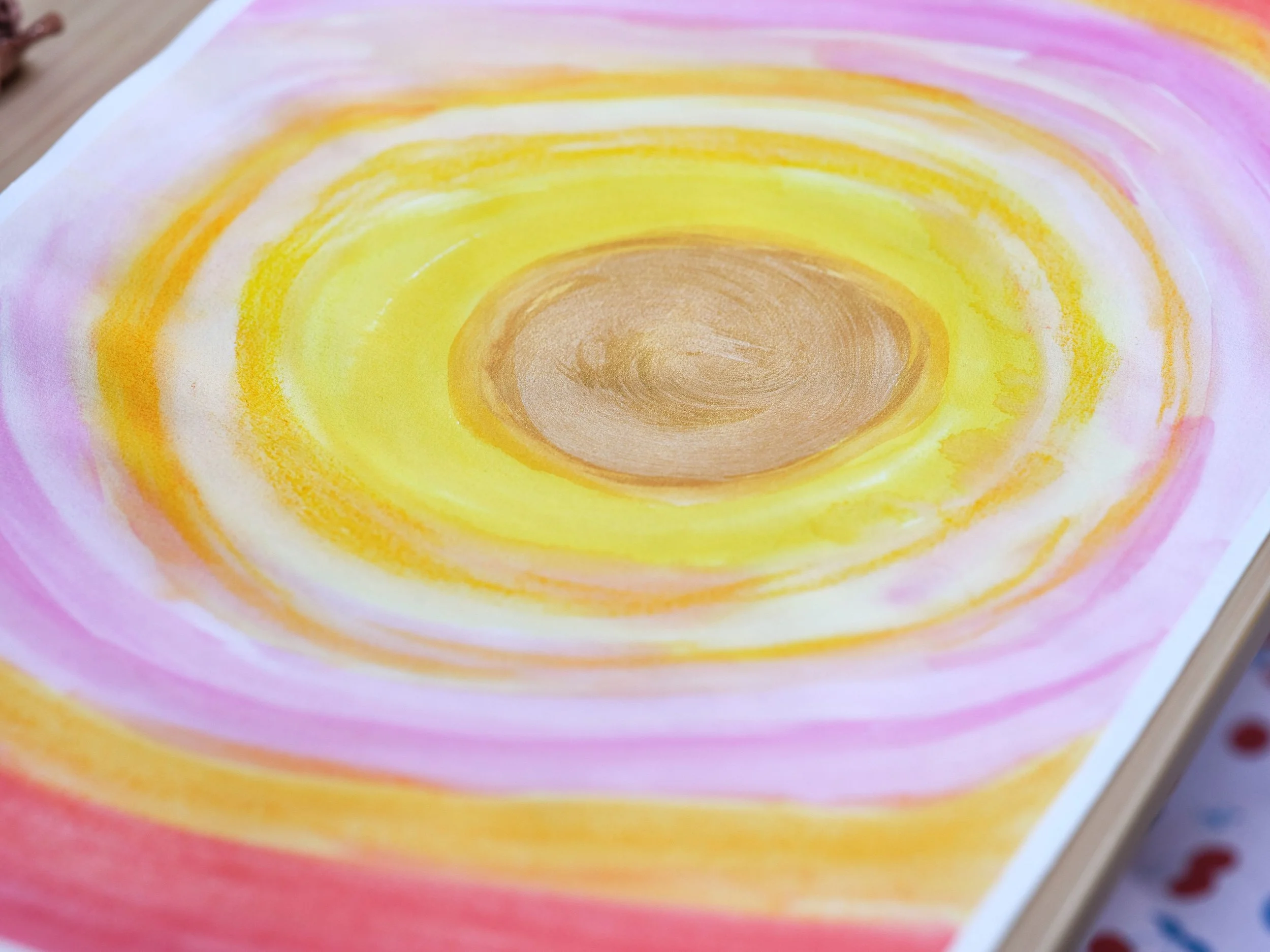 Verwackeltes Bild eines bunten, kreisförmigen Musters mit Farben wie Braun, Gelb, Orange, Rosa und Weiß, das möglicherweise durch Farbstifte oder Pastellkreiden auf Papier erstellt wurde.