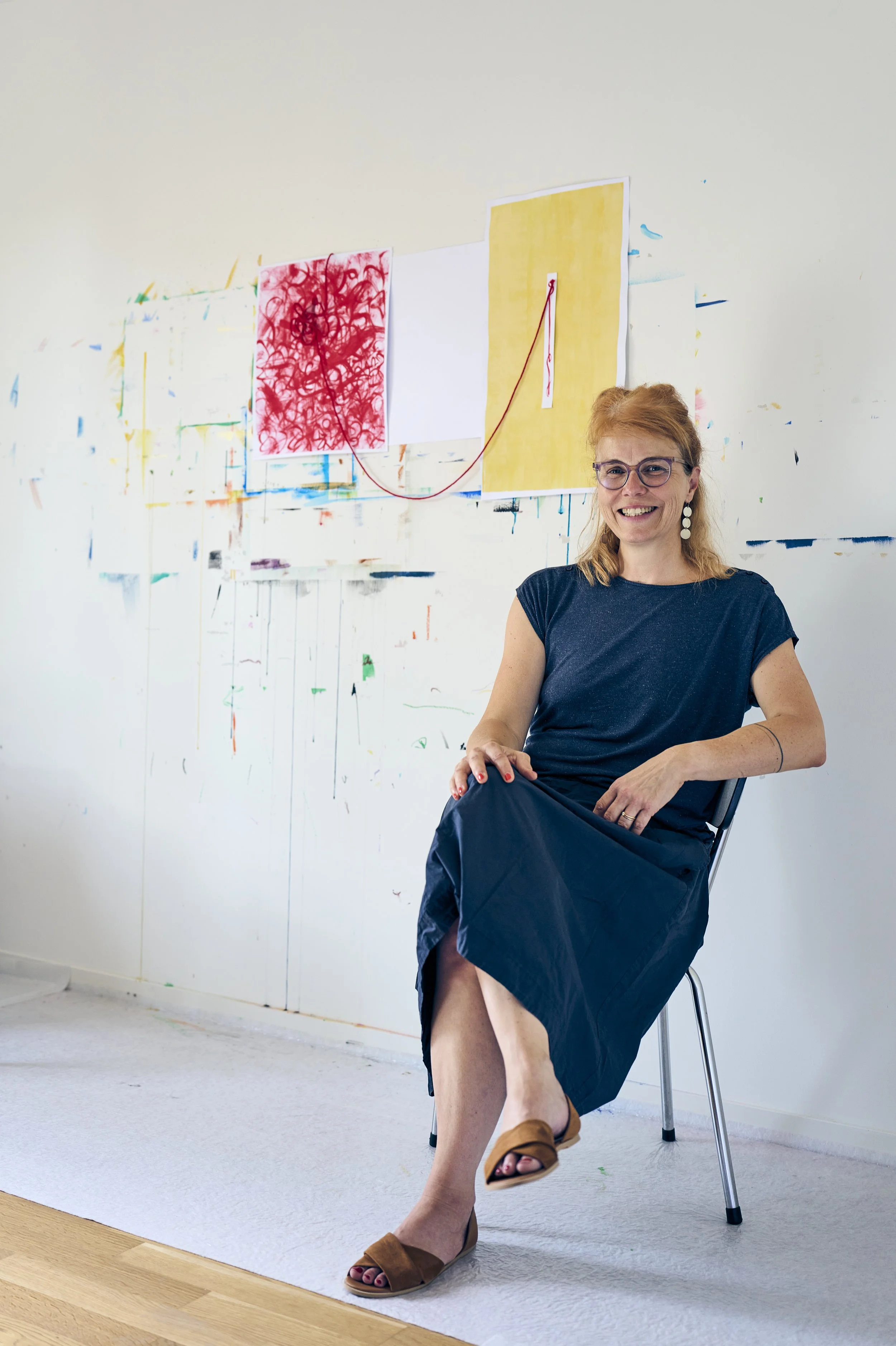 Eine lachende Frau mit roten Haaren, trägt eine schwarze Kleid und braune Schuhe, sitzt auf einem Stuhl vor einer weißen Wand mit bunten Kunstwerken.
