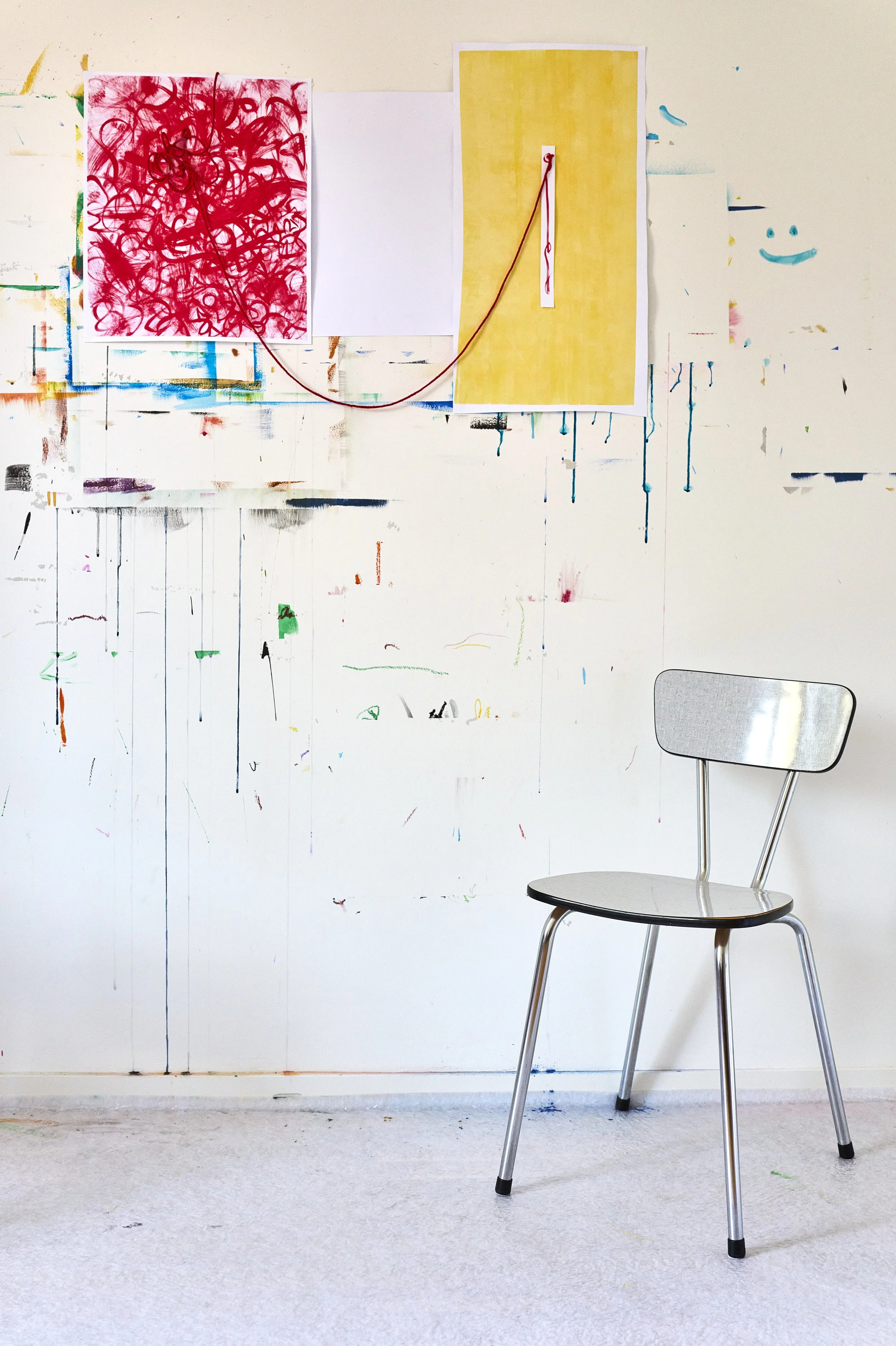 Ein Stuhl steht vor einer weißen Wand, die mit bunten, abstrakten Farbklecksen und Linien bemalt ist. An der Wand sind drei Plakate mit unterschiedlichen Farben und Mustern befestigt, eines rot mit Kringeln, eines weiß, und eines gelb mit einer vertikalen Linie. Das Foto ist in einem Raum aufgenommen, der eine kreative Atmosphäre ausstrahlt.