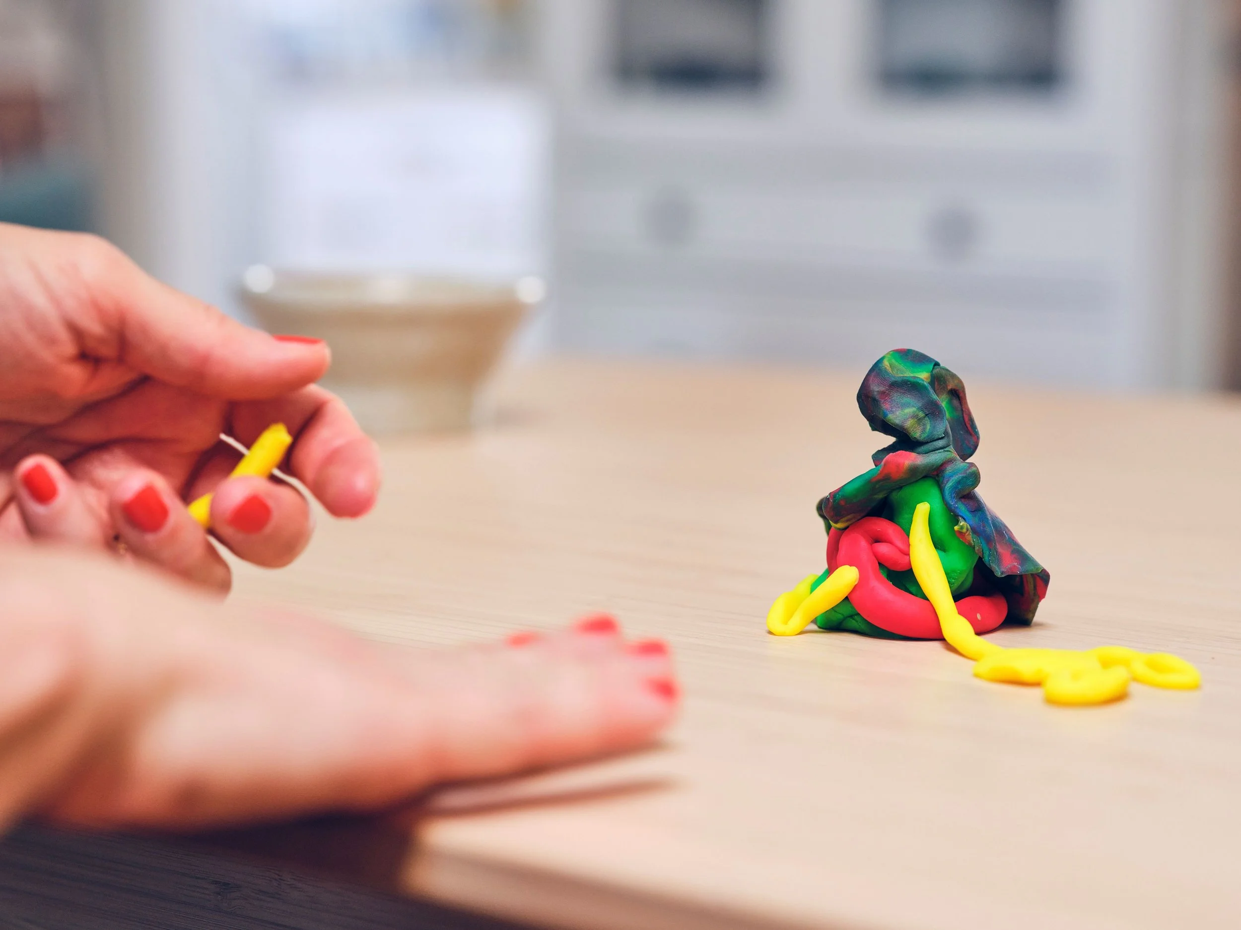 Hand formt eine gelbe Knetfigur, daneben eine bunte Figur aus Knete