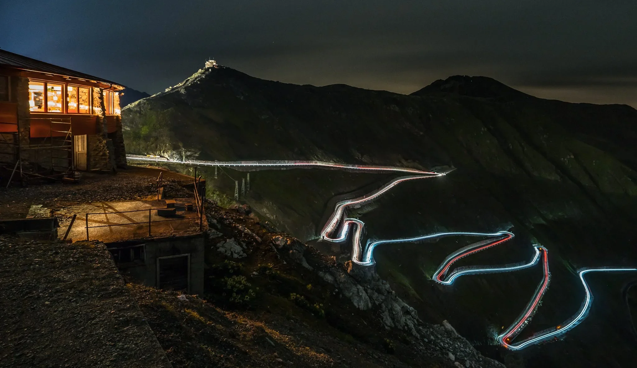 Nighttime photo of a mountain landscape with a winding road illuminated by vehicle lights, a building with illuminated windows on the left, and a mountain peak with a small structure at the top in the background.