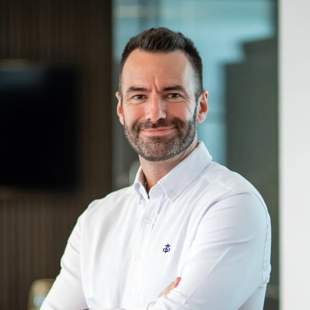 A man with dark hair, a beard, and a mustache, wearing a white button-up shirt, smiling with crossed arms in an indoor office environment.