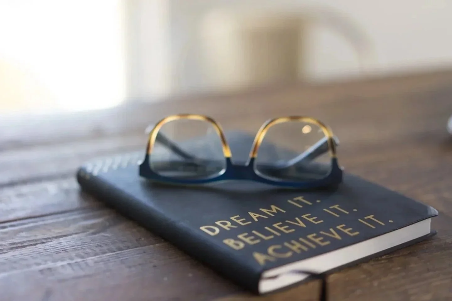 Eyeglasses resting on a notebook with the motivational quote 'DREAM IT. BELIEVE IT. ACHIEVE IT.' on a wooden surface.