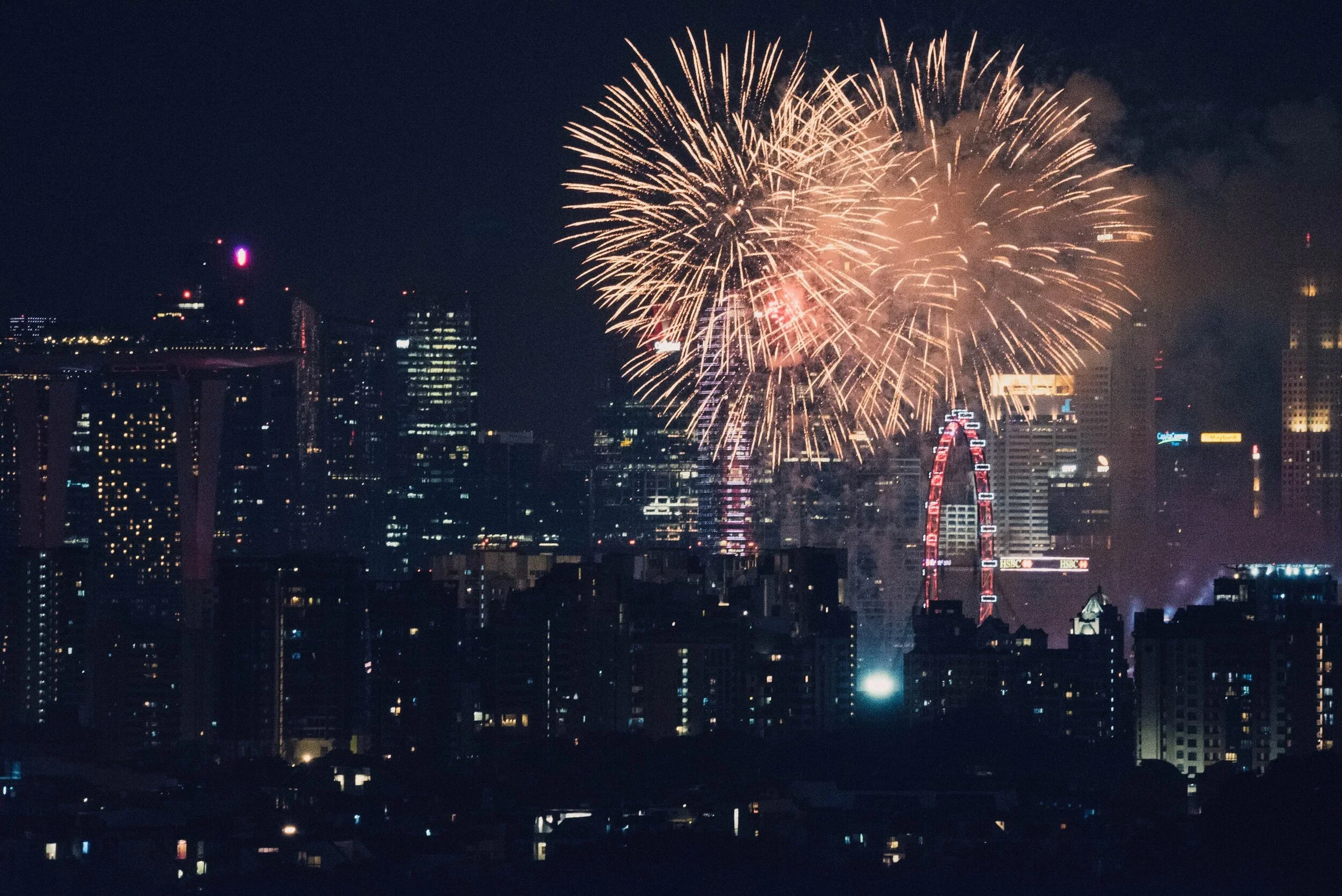 Nighttime city skyline with fireworks illuminating the sky, including an iconic ferris wheel.