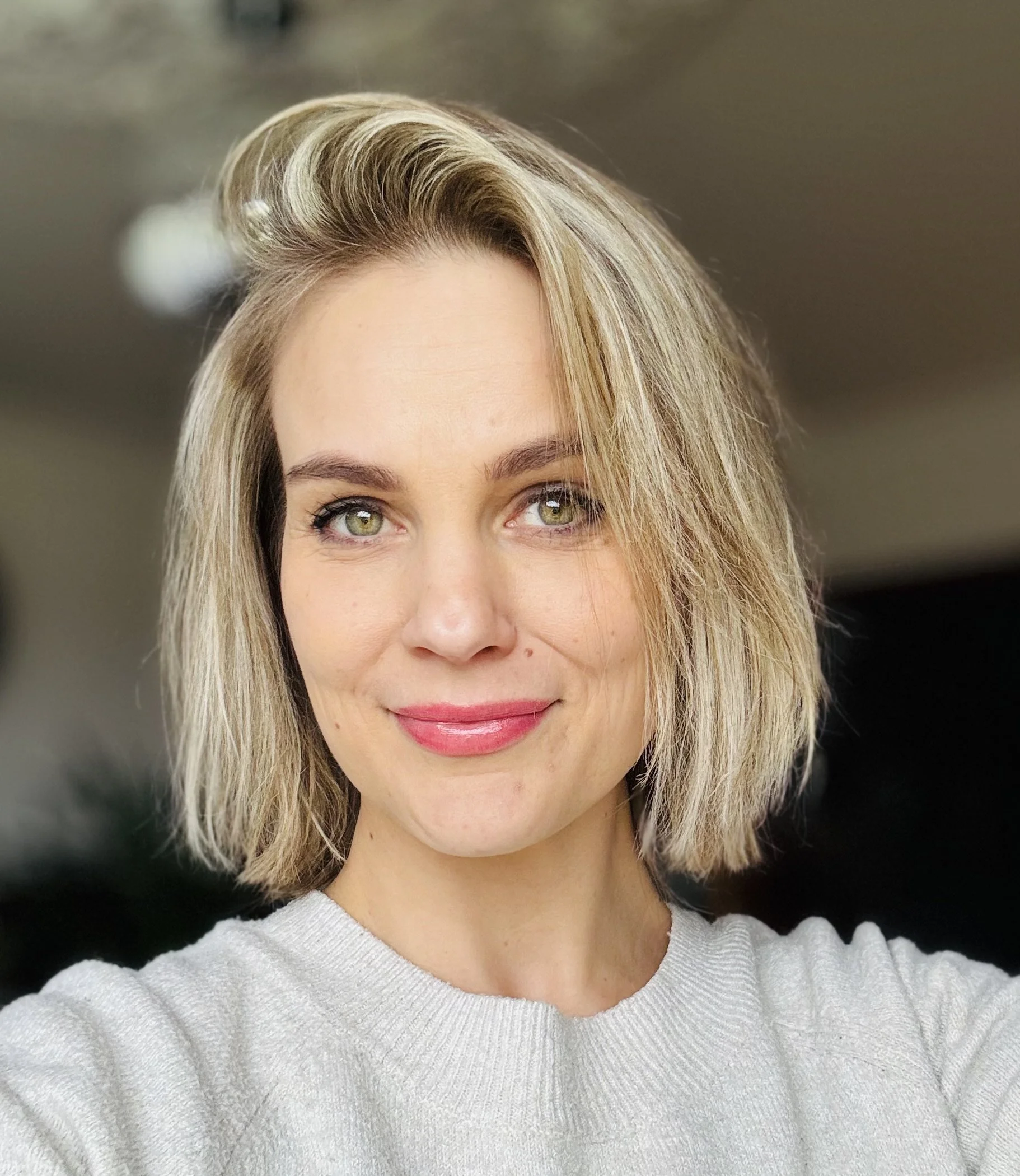 Nahaufnahme einer lachenden Frau mit blonden, welligen Haaren und grünen Augen, die einen hellen Pullover trägt.