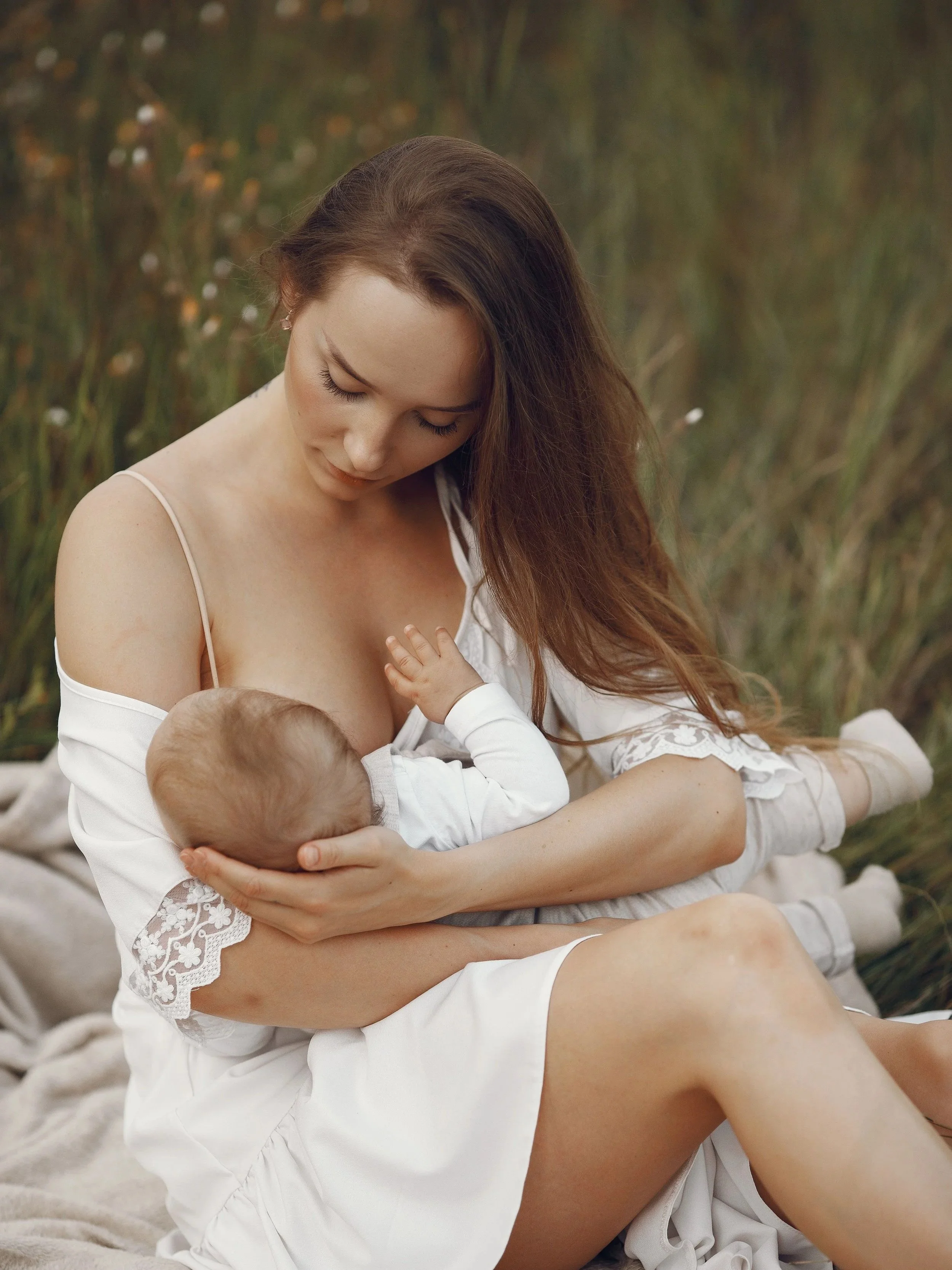 Une femme avec des cheveux longs et bruns en tenue blanche, tenant un bébé blond dans ses bras, assise sur un tapis beige, avec des plantes vertes en arrière-plan.