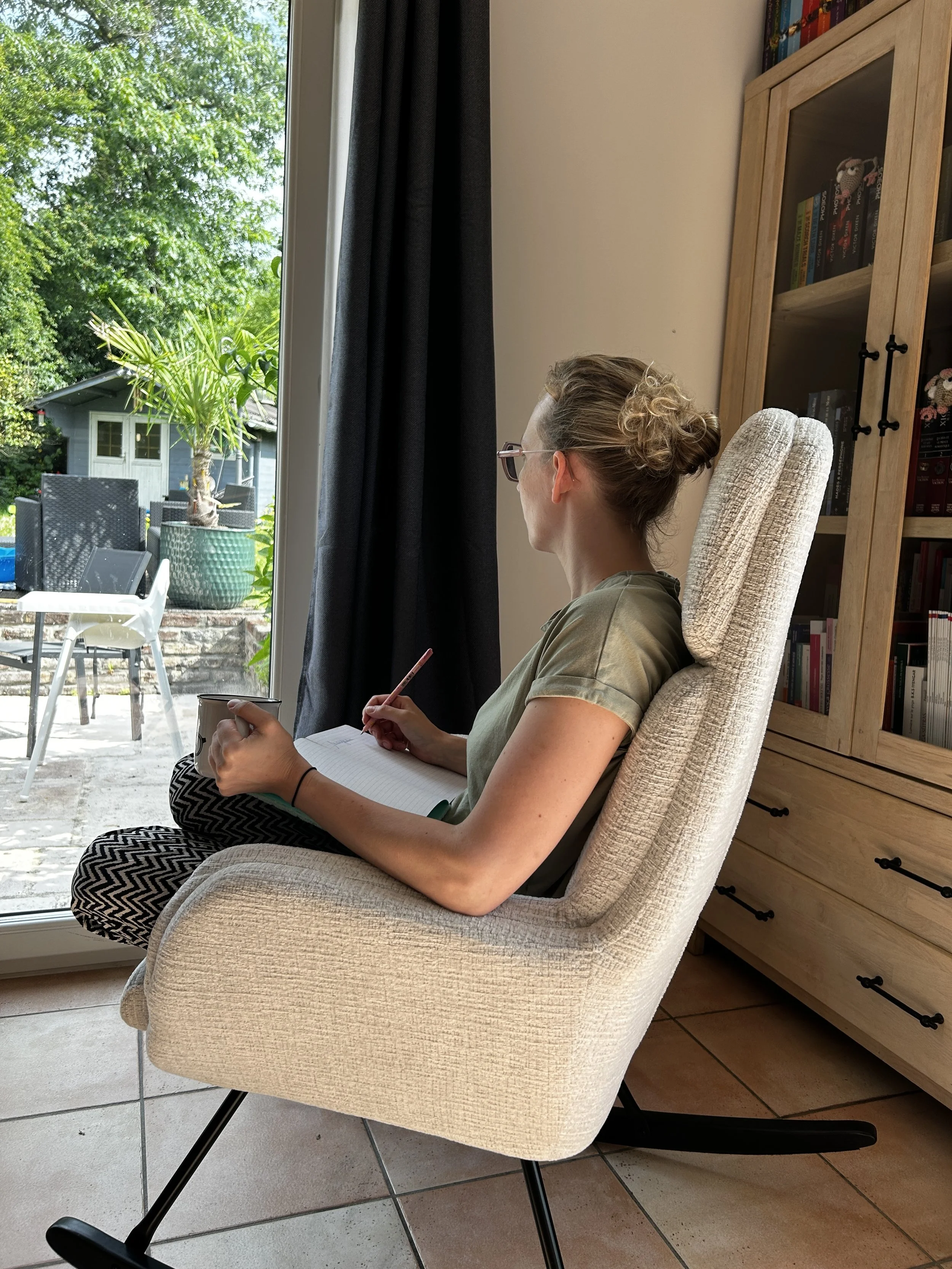 Femme assise dans un fauteuil beige, regardant par une fenêtre ouverte vers un jardin avec un arbre et des meubles d'extérieur, tenant un mug et un carnet, portant des lunettes et des vêtements verts.