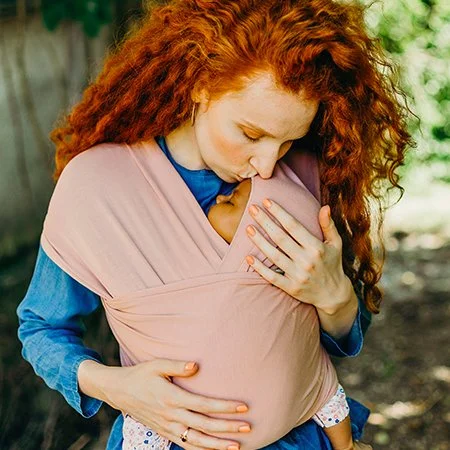 Une femme avec des cheveux roux porte un bébé dans une écharpe de portage, elle regarde l'enfant avec tendresse dans un cadre extérieur.