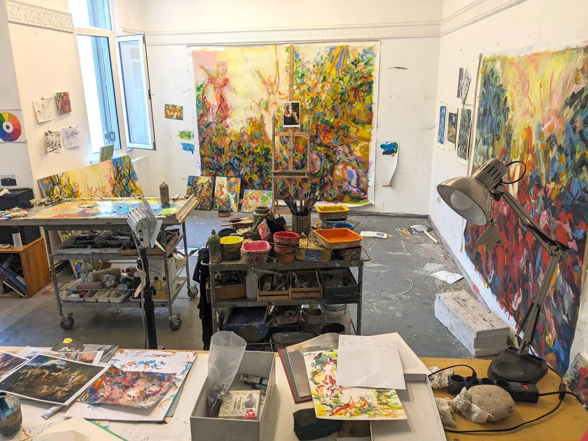 An artist's studio filled with colorful paintings, brushes, and art supplies. Large finished paintings hang on the walls, featuring vibrant, abstract, and figurative styles, with some paintings leaning against the wall.