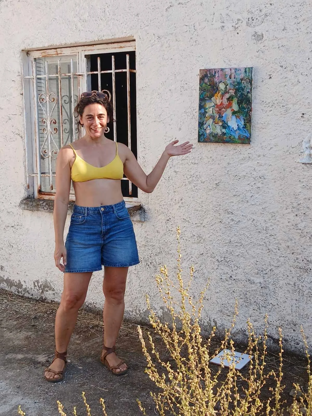 A woman in a yellow crop top and denim shorts standing outside in front of a textured off-white wall with an art piece hanging on it. She is smiling and gesturing towards the artwork.