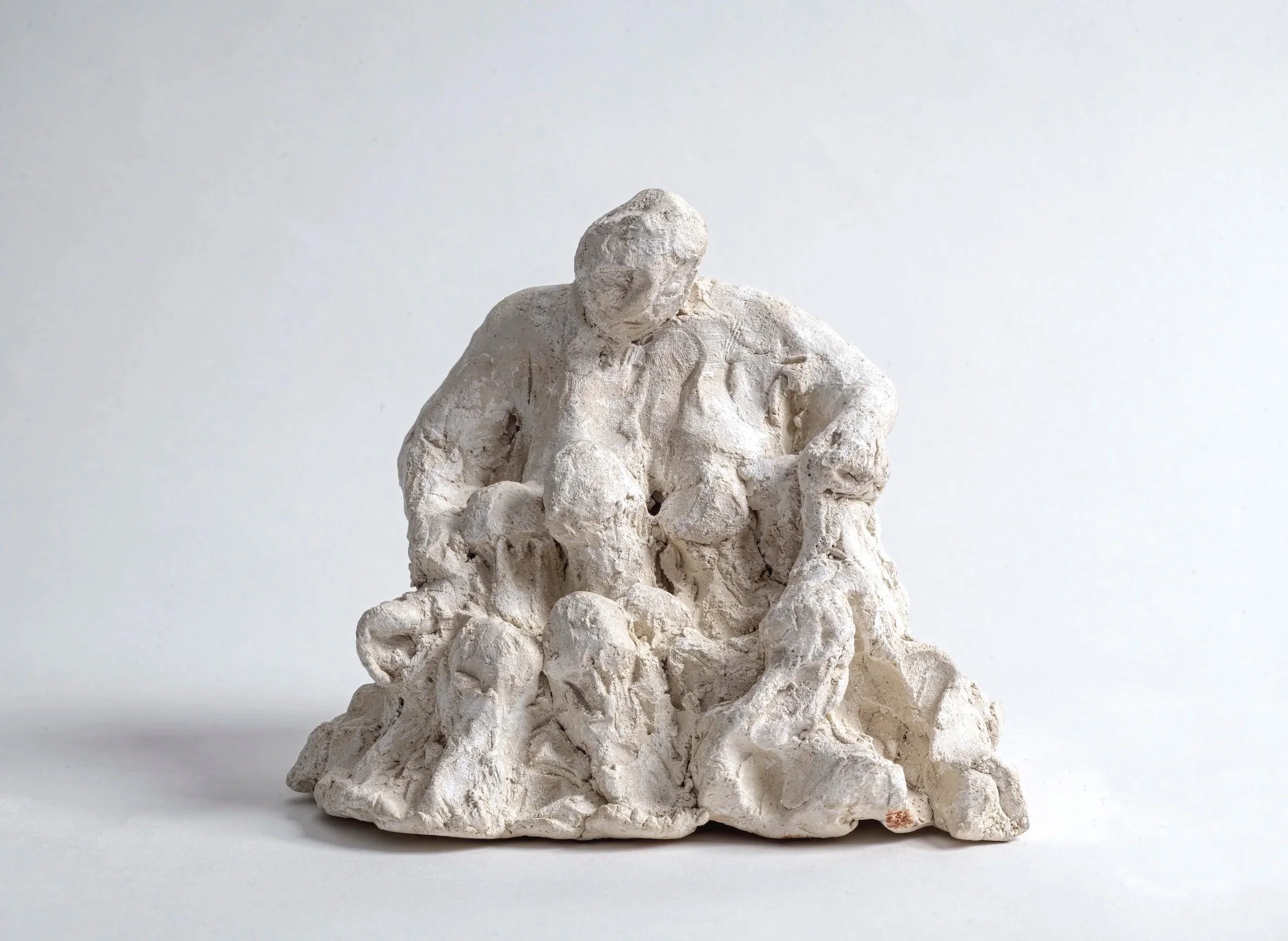 White sculpture of a seated man with a bearded face, dressed in a coat, placed on a rough, uneven base, against a plain white background.