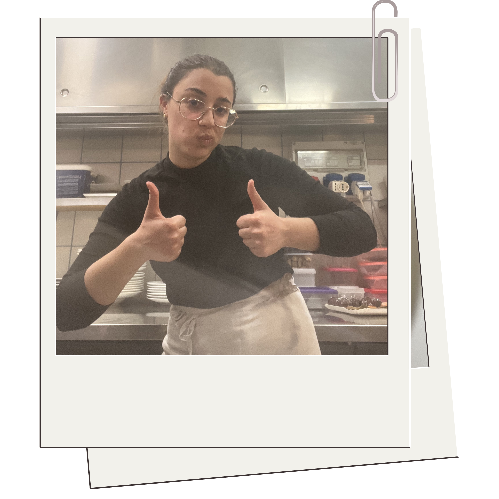 A young woman with glasses giving a thumbs up while standing in a kitchen, behind her are kitchen appliances and utensils.